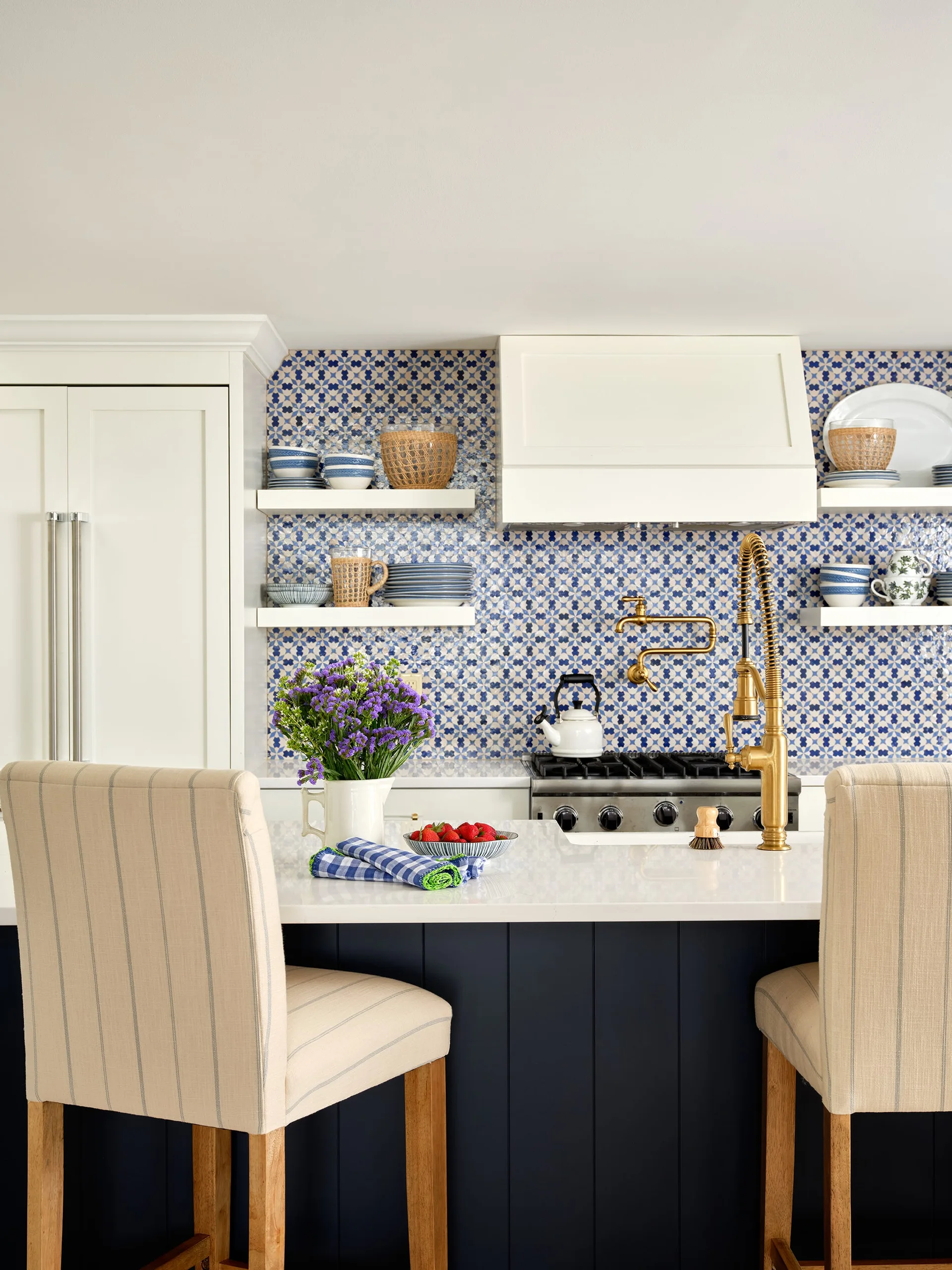 A modern transitional kitchen featuring a navy blue island, white countertops, striped bar stools, and a blue and white patterned tile backsplash.