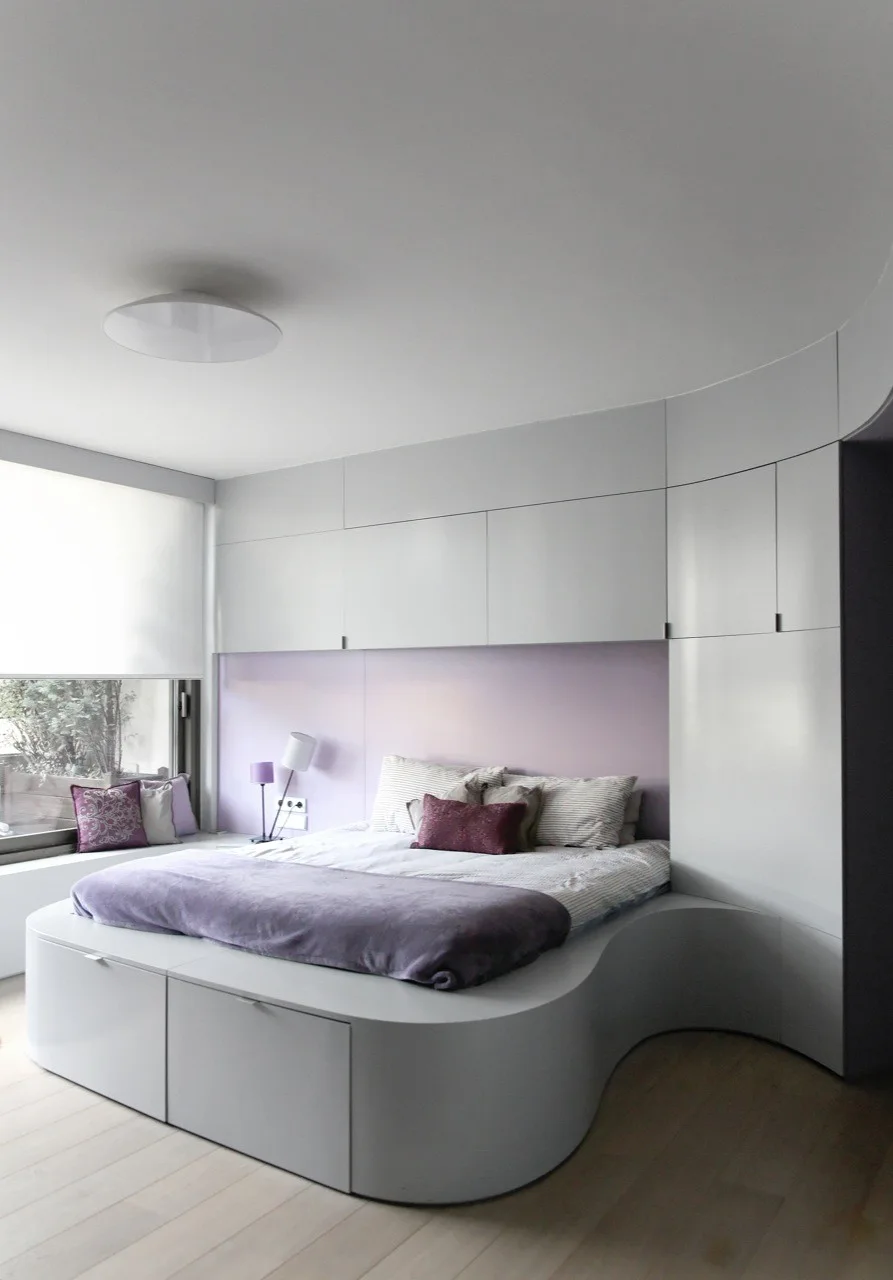 A modern bedroom with a custom curved white platform bed integrated with overhead wall cabinets against a soft lavender backdrop panel.