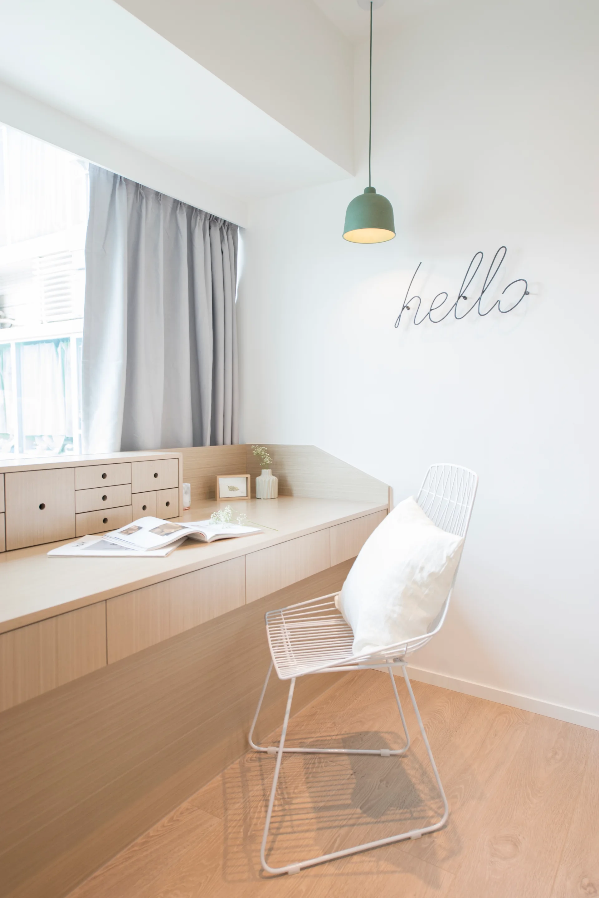 A minimalist home office features a light wood built-in desk, a white wire chair with a pillow, and a sage green pendant light against a white wall.
