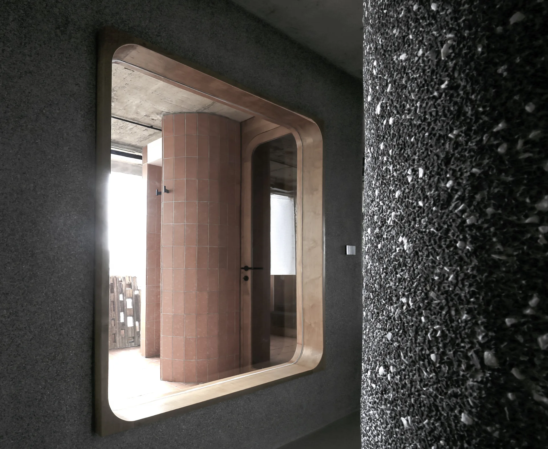 A rounded wooden frame set in a textured grey concrete wall looks through to a curved terracotta tiled wall.
