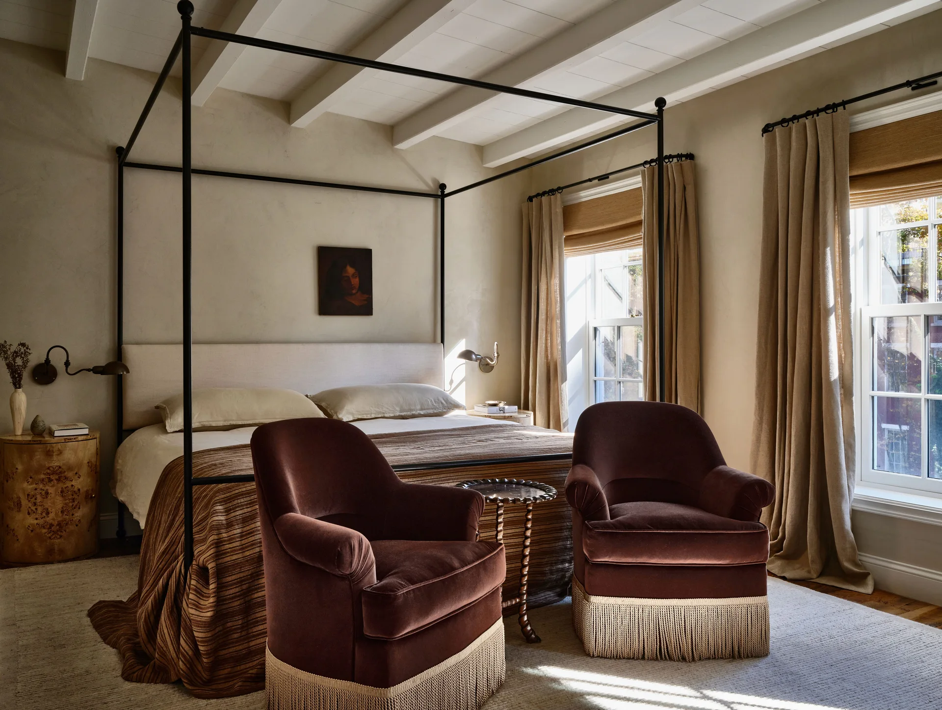 A bedroom featuring a black metal canopy bed and two brown velvet armchairs with fringe trim on a cream-colored rug.