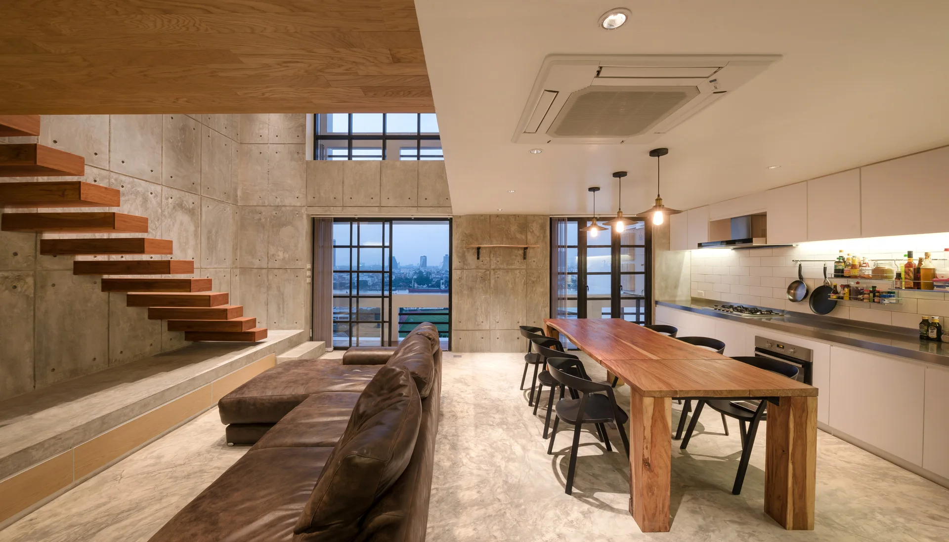 An open loft apartment featuring a floating wooden staircase against an exposed concrete wall beside a long dining table.