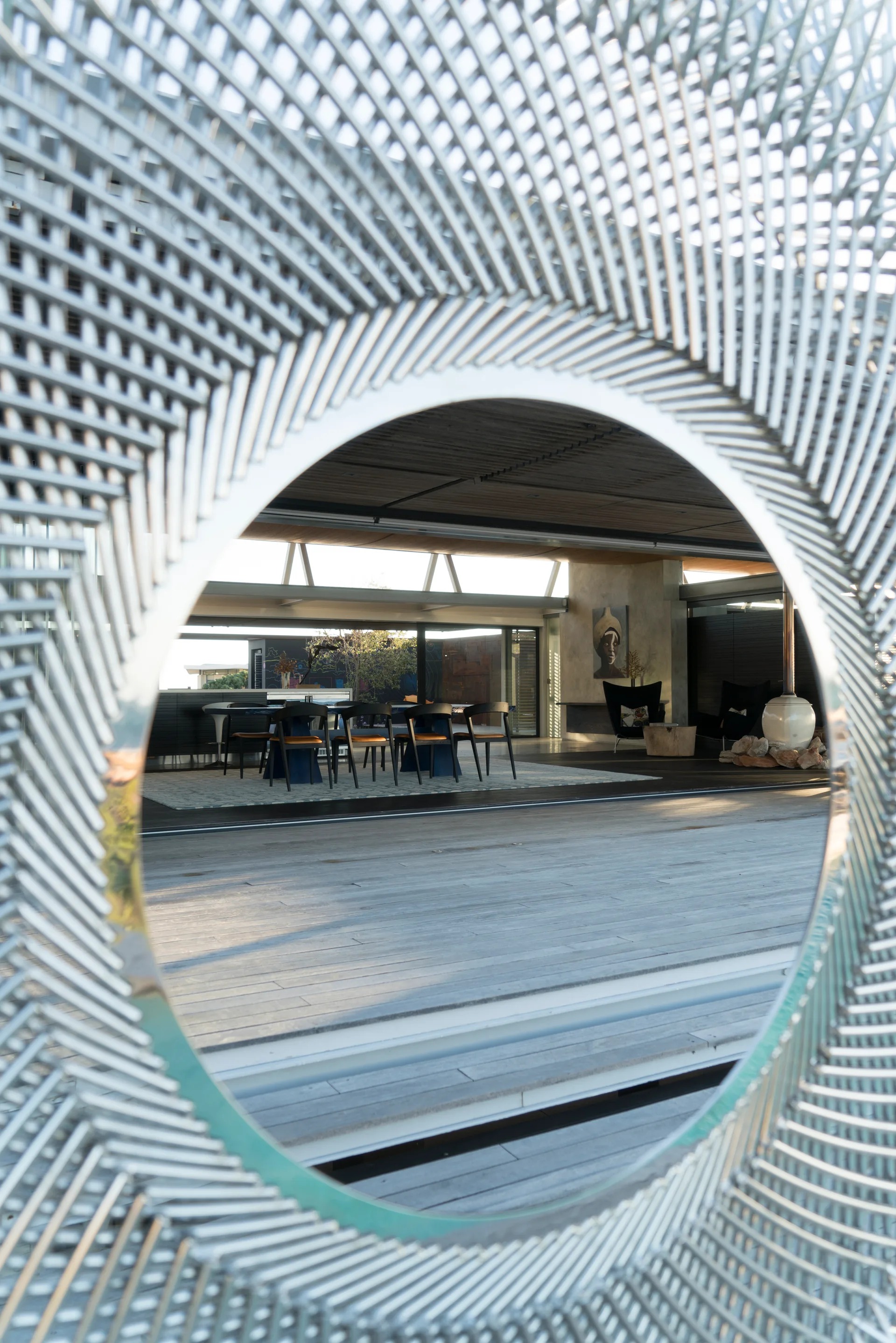 A circular patterned metal screen frames a view of a covered outdoor dining patio with a table, black chairs, and wood plank flooring.
