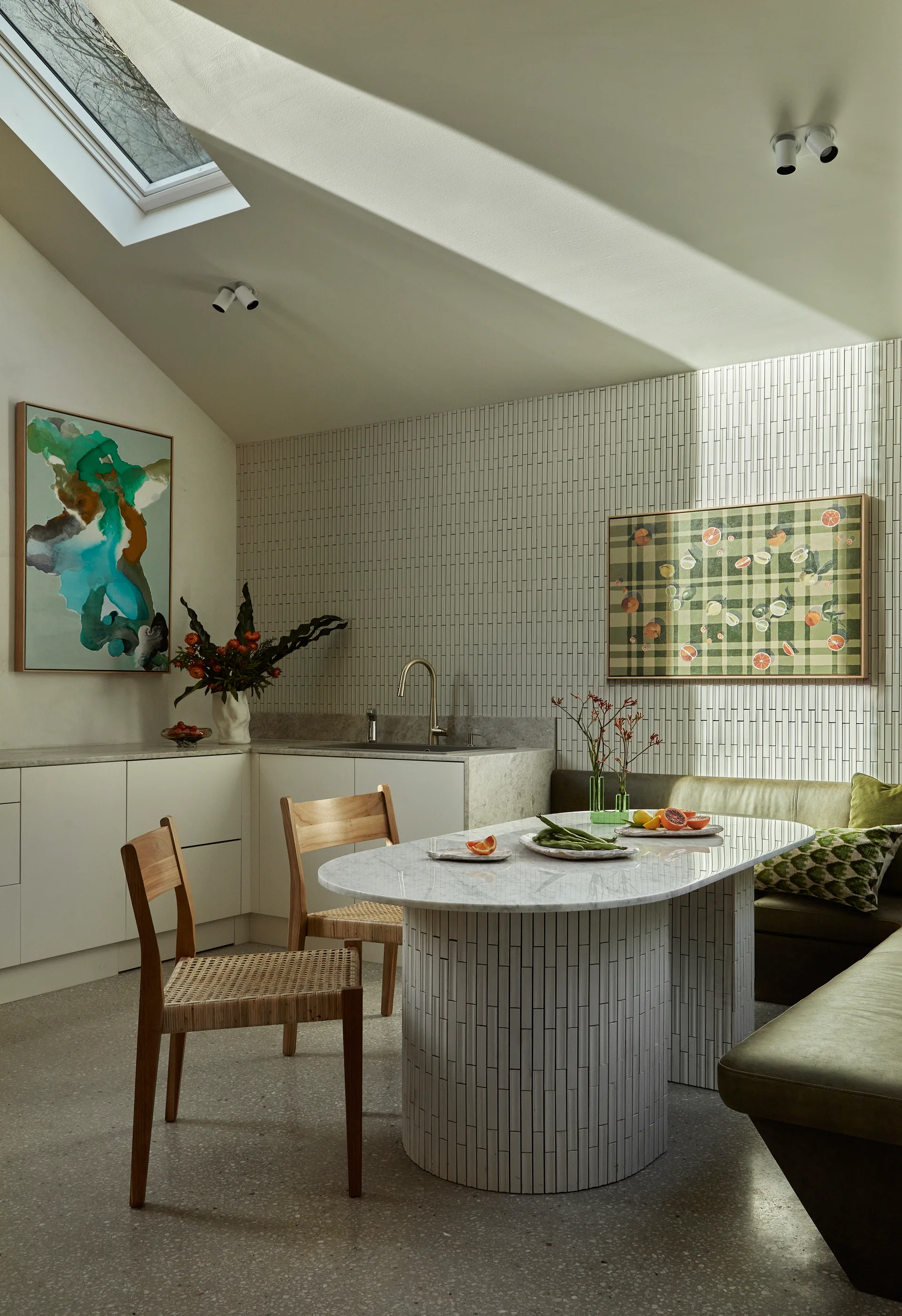 A dining nook features an oval marble table with a tiled base, wooden chairs, a green built-in bench, and a vertically tiled accent wall.