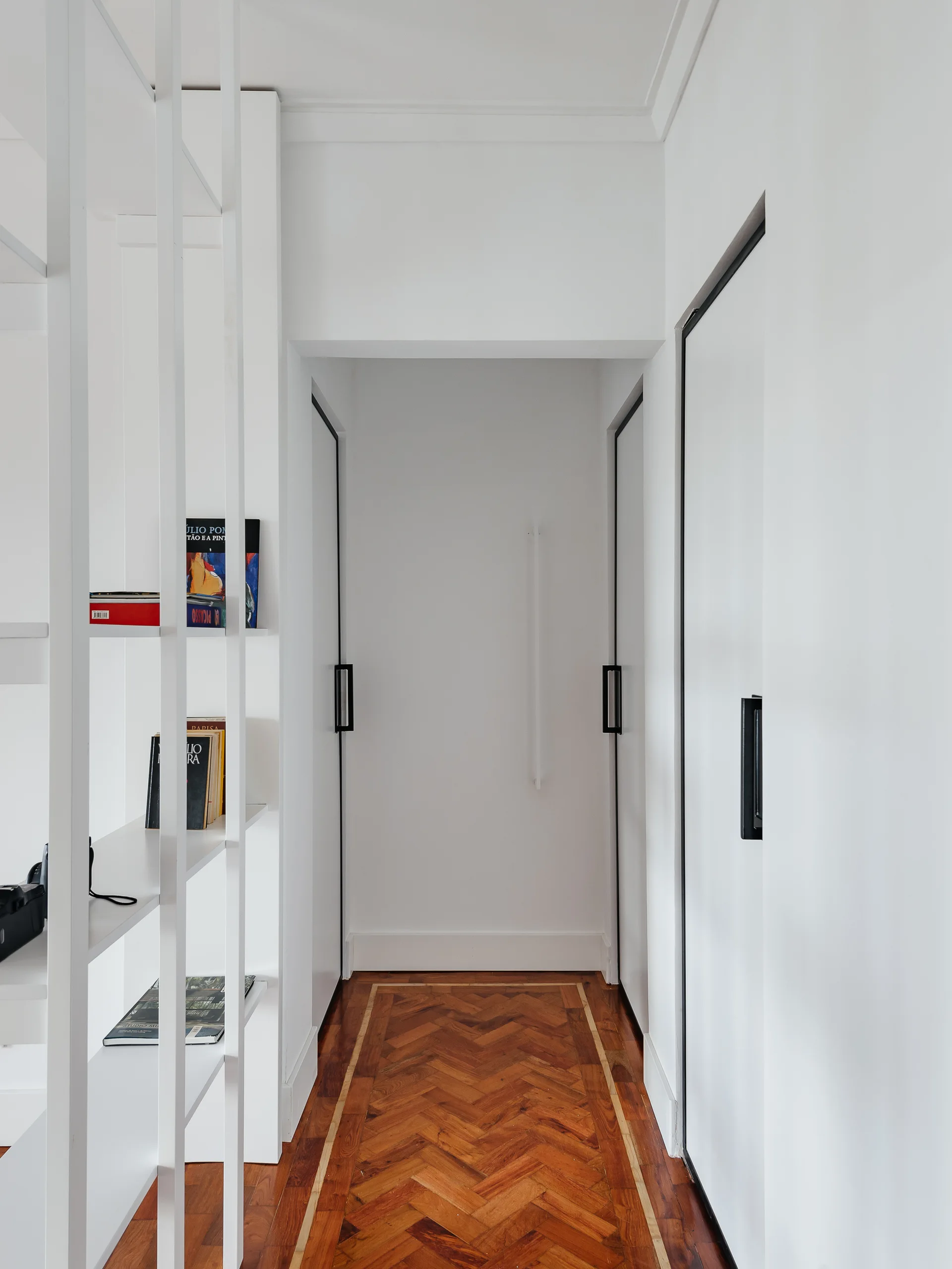 A narrow white hallway with a rich brown herringbone parquet floor, white flush doors with black handles, and a white open shelving frame.