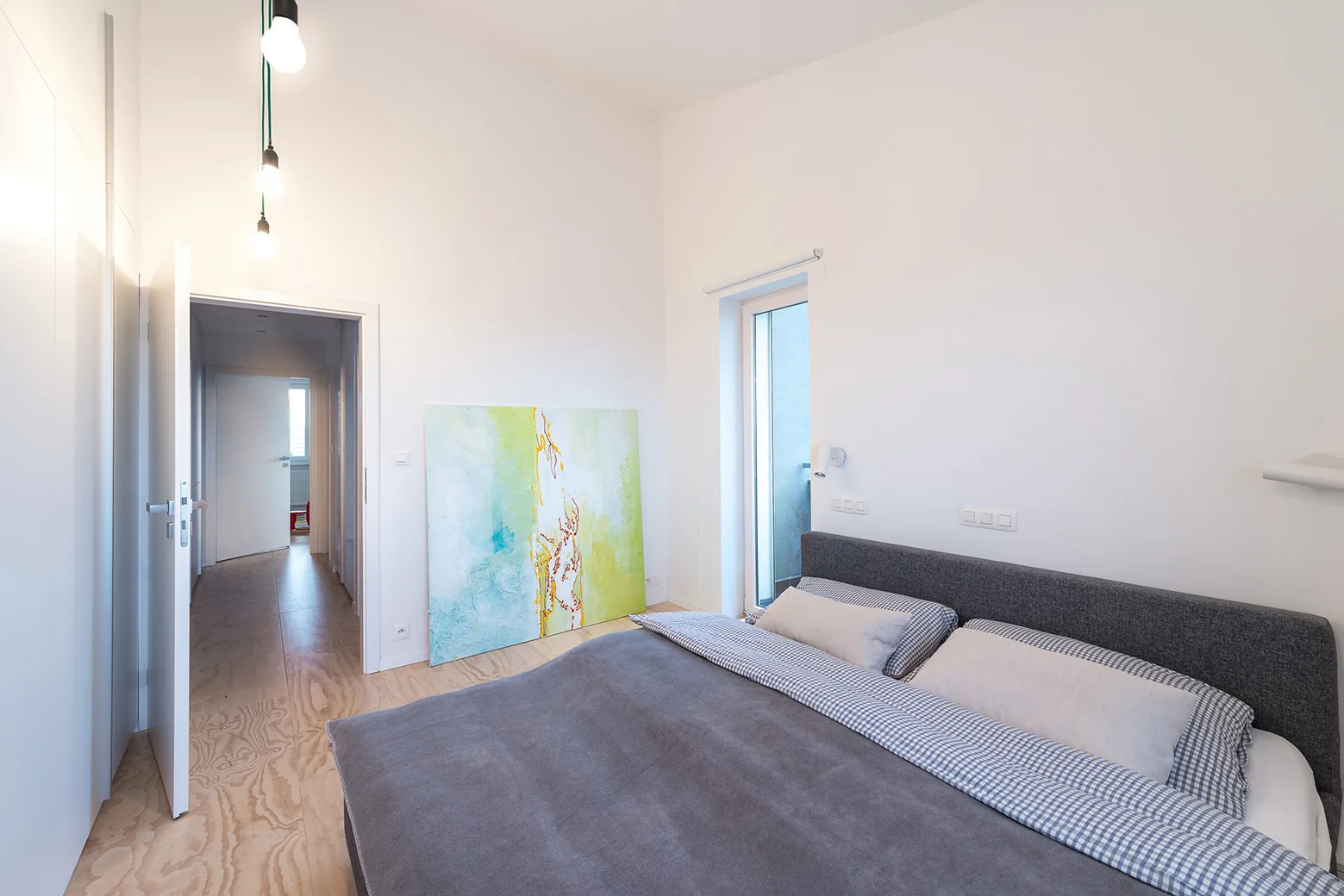 A minimalist white bedroom featuring a grey upholstered bed with checkered bedding, plywood flooring, and a large abstract painting on the floor.