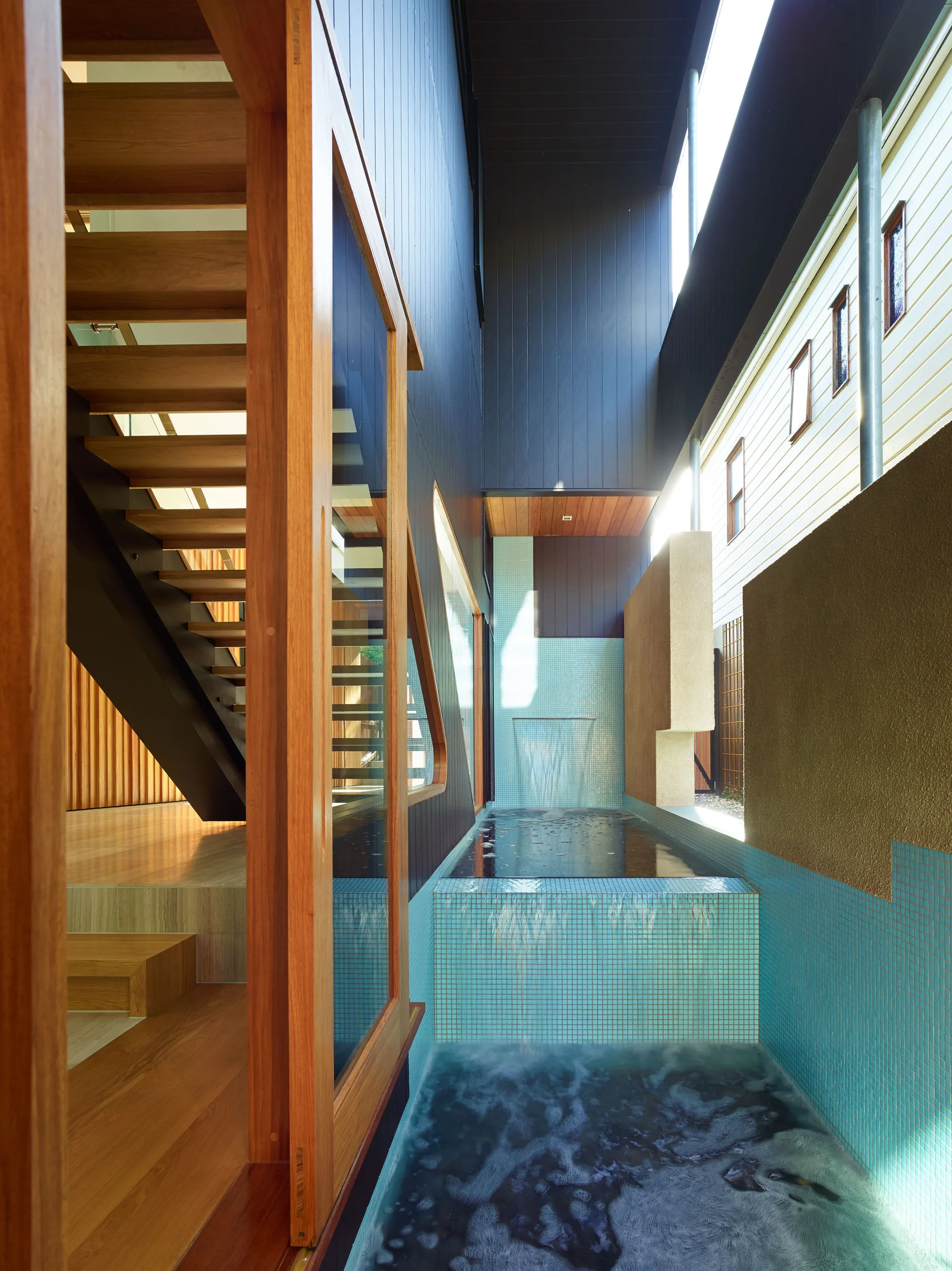 A narrow courtyard featuring a multi-tiered pool with bright blue mosaic tiles next to a modern wooden staircase visible through large windows.