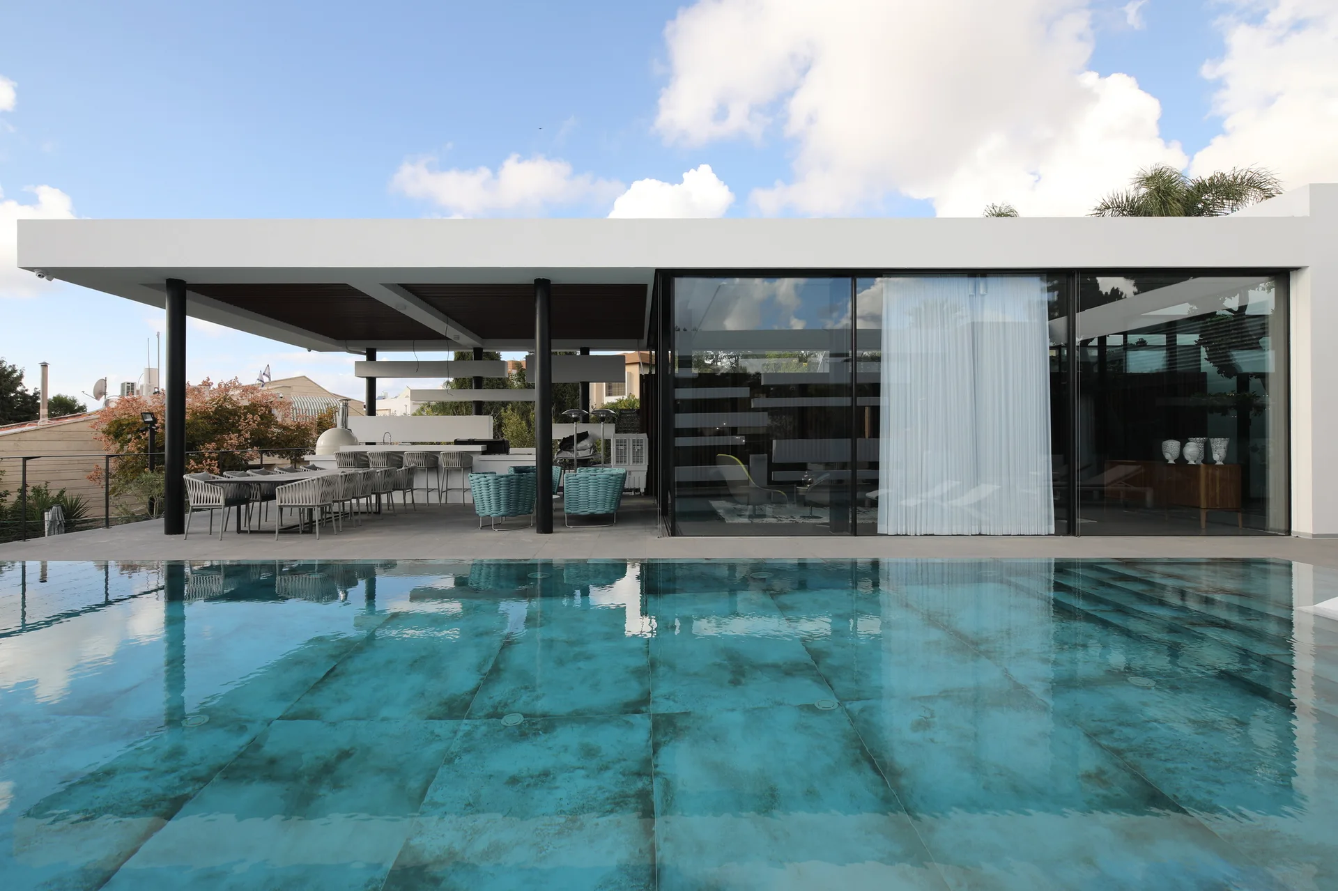 A contemporary swimming pool with turquoise tiled water sits adjacent to a covered patio featuring an outdoor kitchen and large glass-walled living spaces.