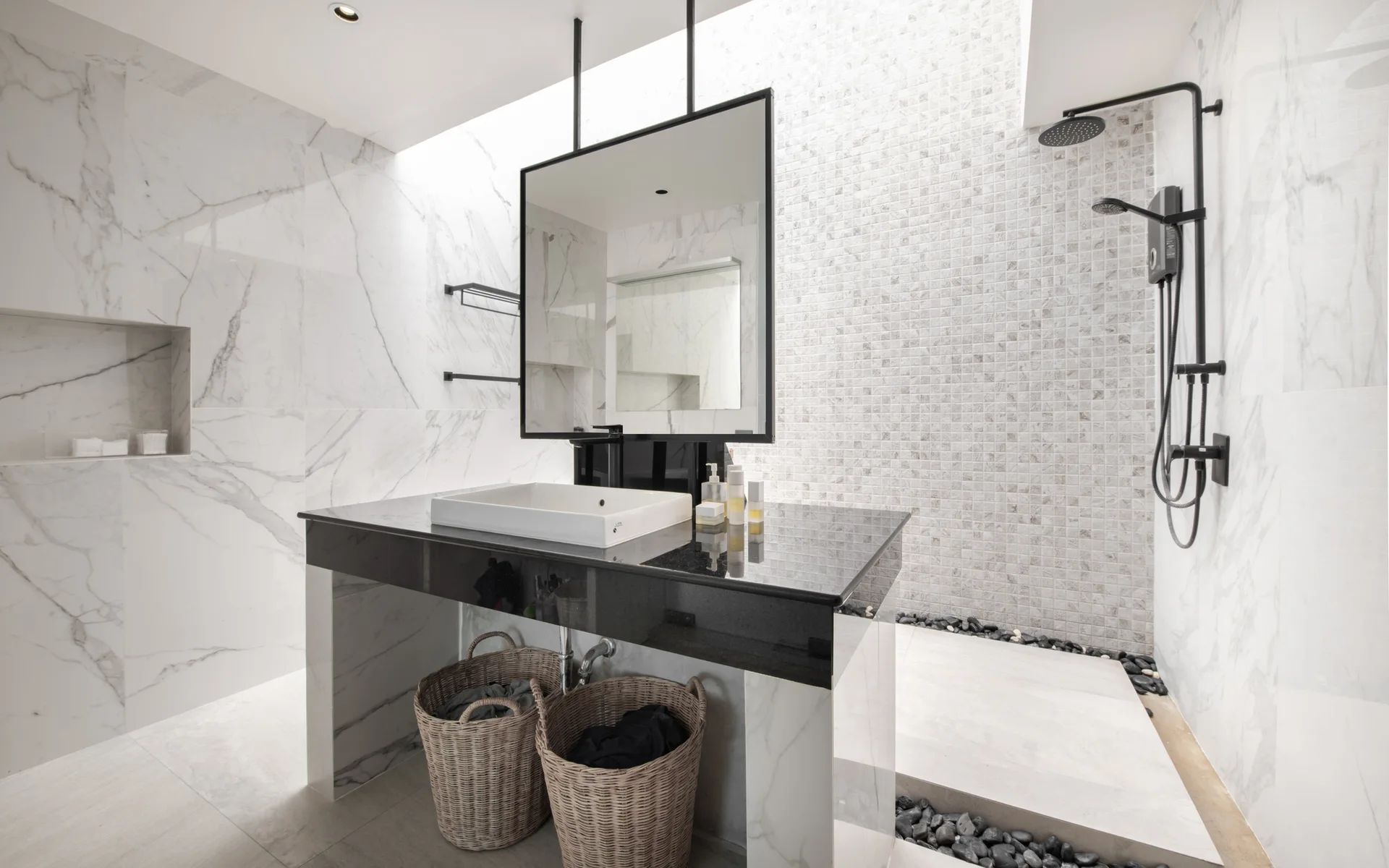 A modern marble bathroom featuring a black stone vanity under a ceiling-hung mirror, a skylight, and a shower lined with black pebbles.