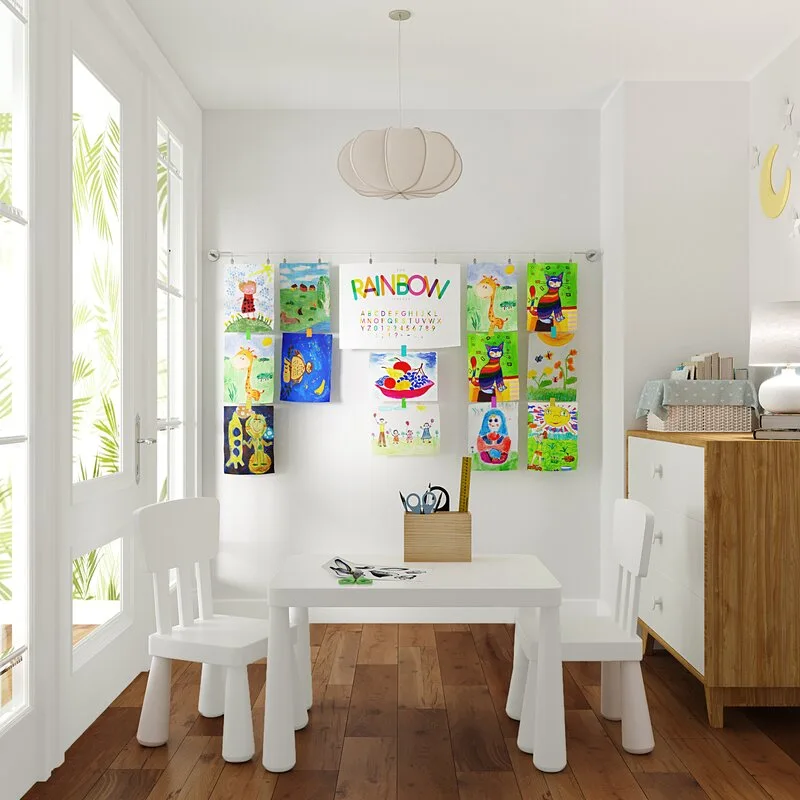 A child-sized white play table and two chairs sit on a wood floor in a playroom featuring colorful drawings clipped to a wall-mounted wire.