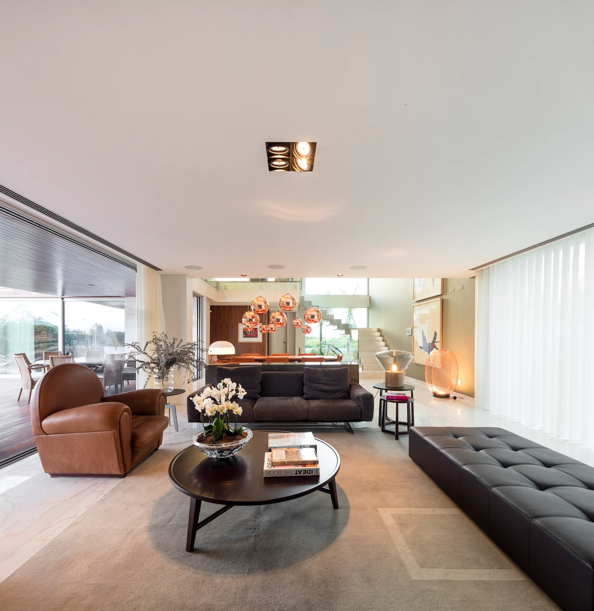 A contemporary open-plan living room with a dark sofa, tan leather armchair, round coffee table, and a black tufted leather bench on a beige rug.