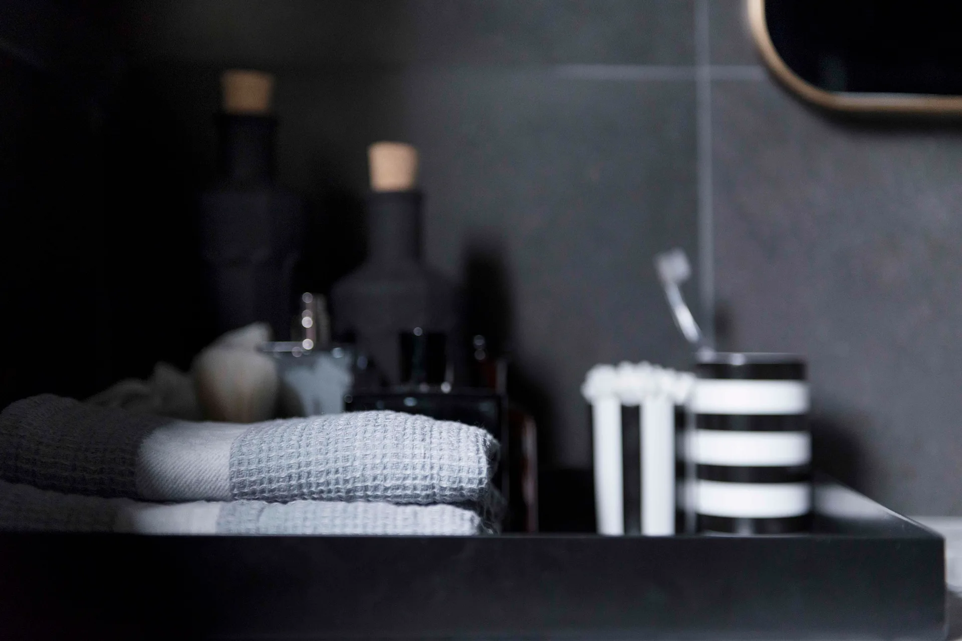 A close-up of a black bathroom vanity tray holding a folded gray waffle weave towel and striped containers against a dark tiled wall.
