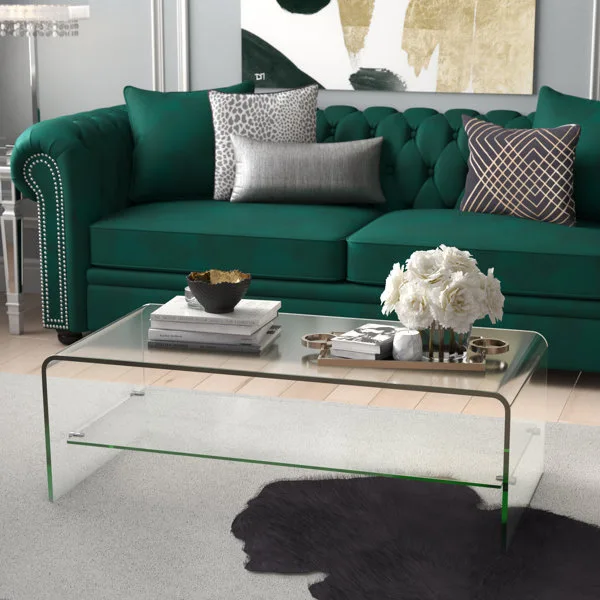 A living room features an emerald green velvet chesterfield sofa paired with a clear glass waterfall coffee table over a grey rug.