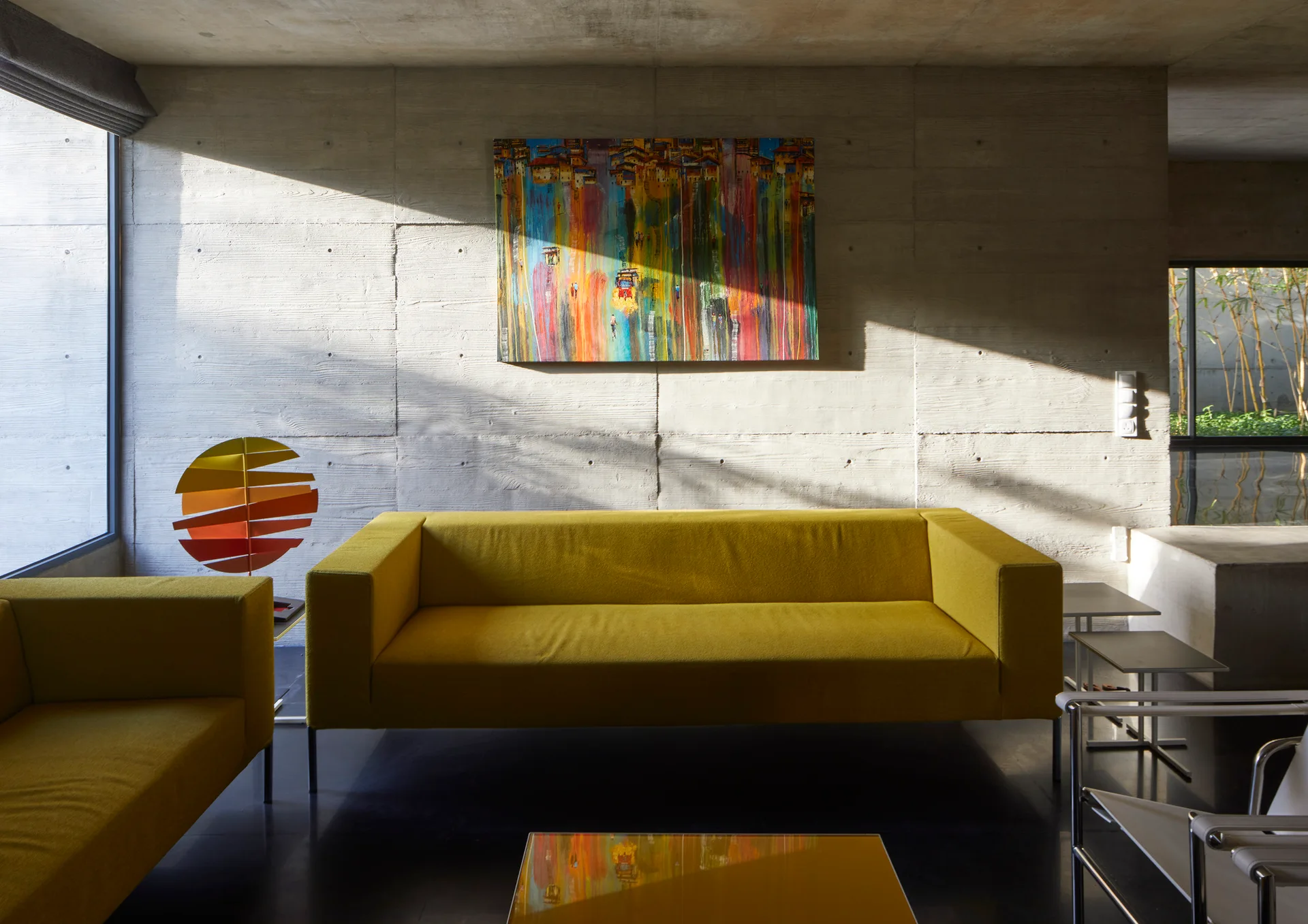 A modern living room features two yellow sofas, a colorful abstract painting, and a raw board-marked concrete wall under bright sunlight.