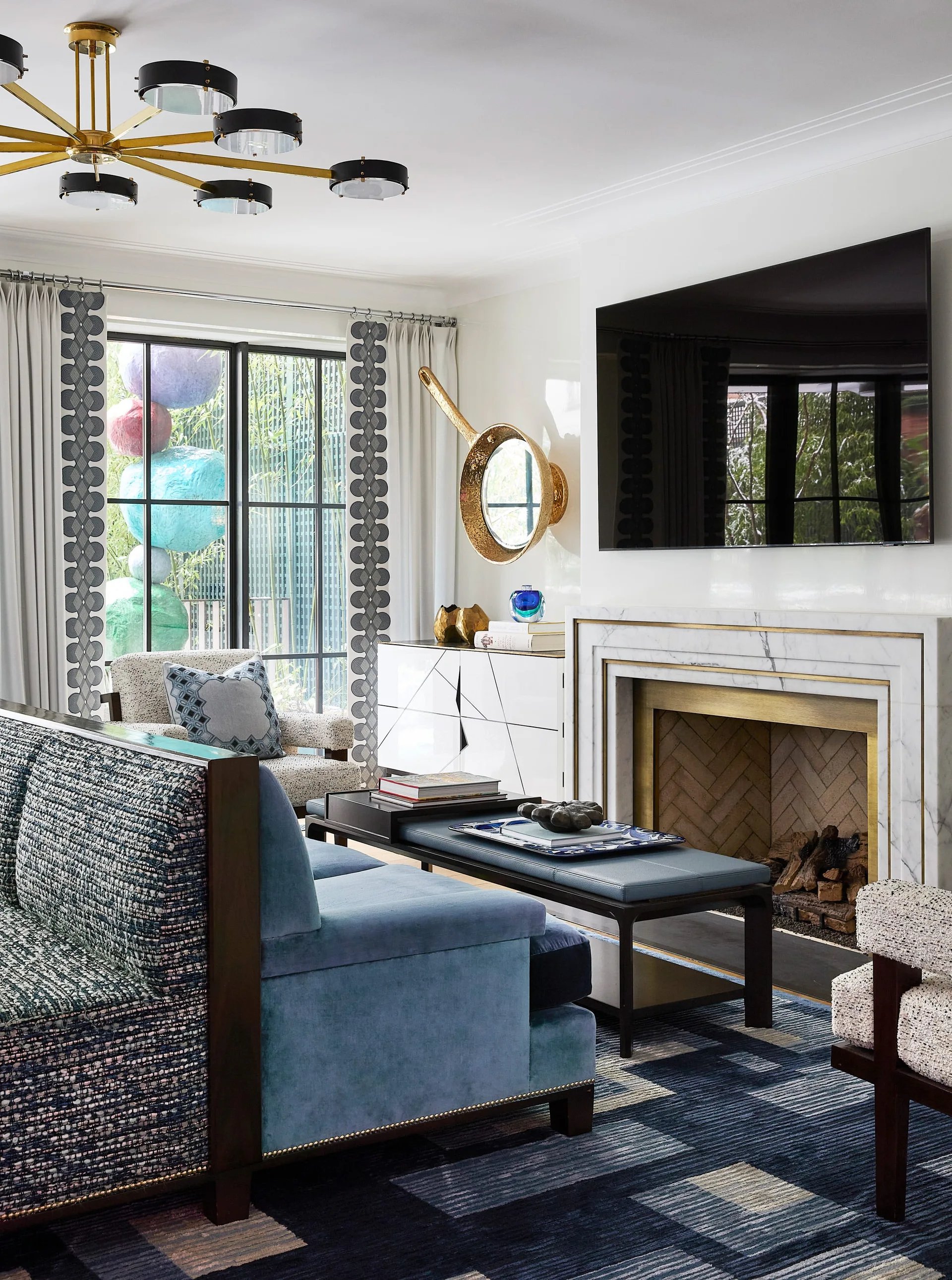 A modern living room featuring a blue velvet sofa, a white marble-surround fireplace with gold trim, a television, and a patterned area rug.