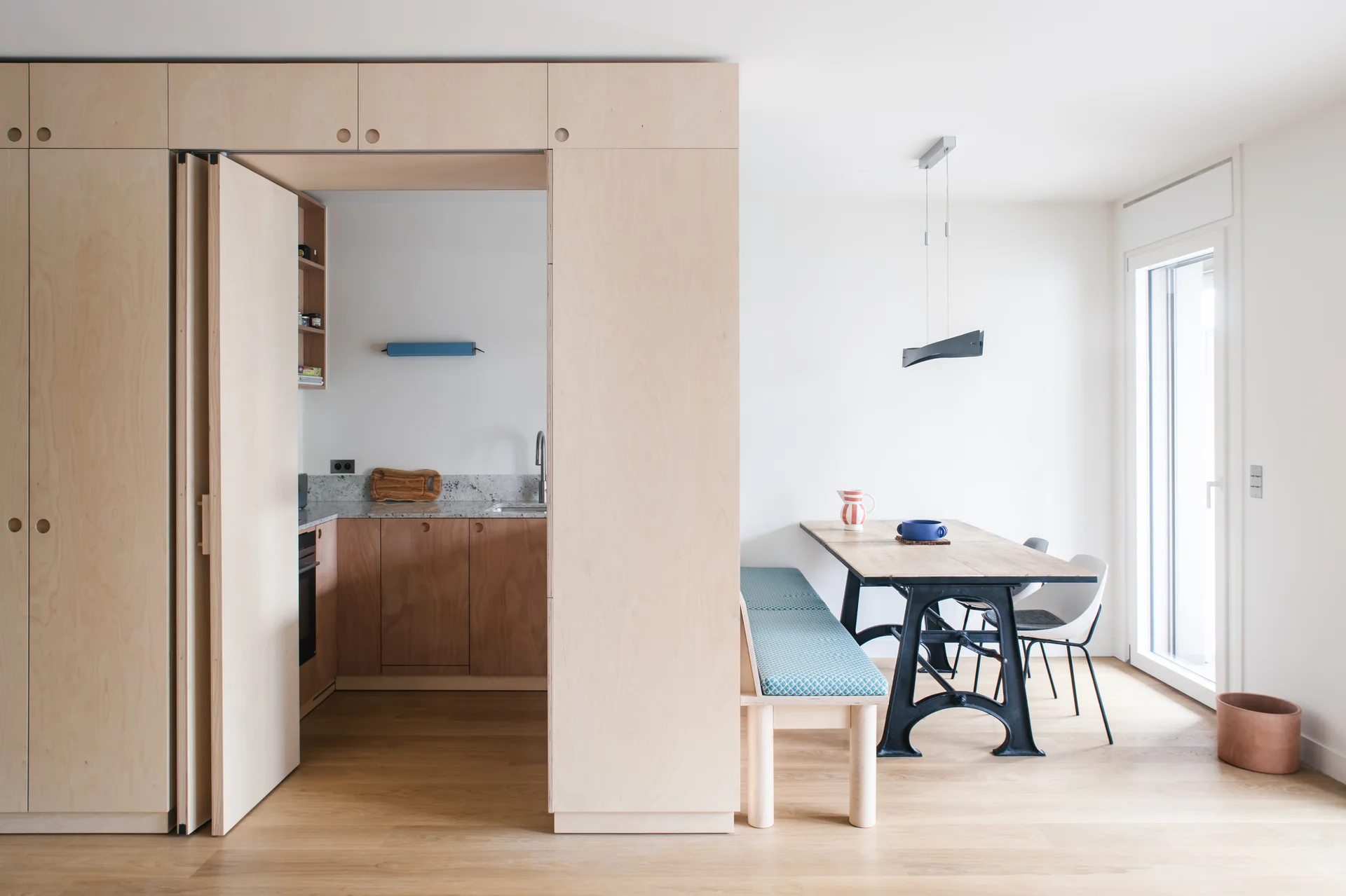A light-filled dining room features a wooden table with a blue cushioned bench next to a large plywood wall that opens to a concealed kitchen.