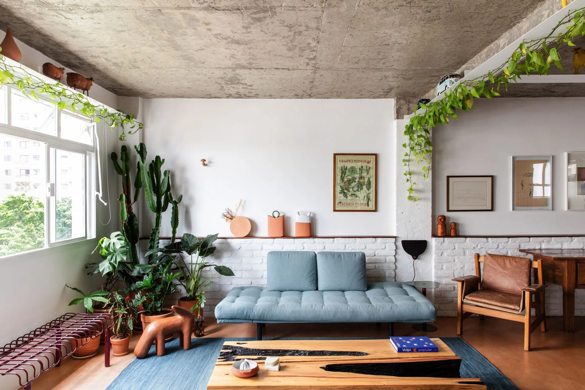 An eclectic living room features a blue daybed sofa on a blue rug, set against a white brick wall below a raw exposed concrete ceiling.
