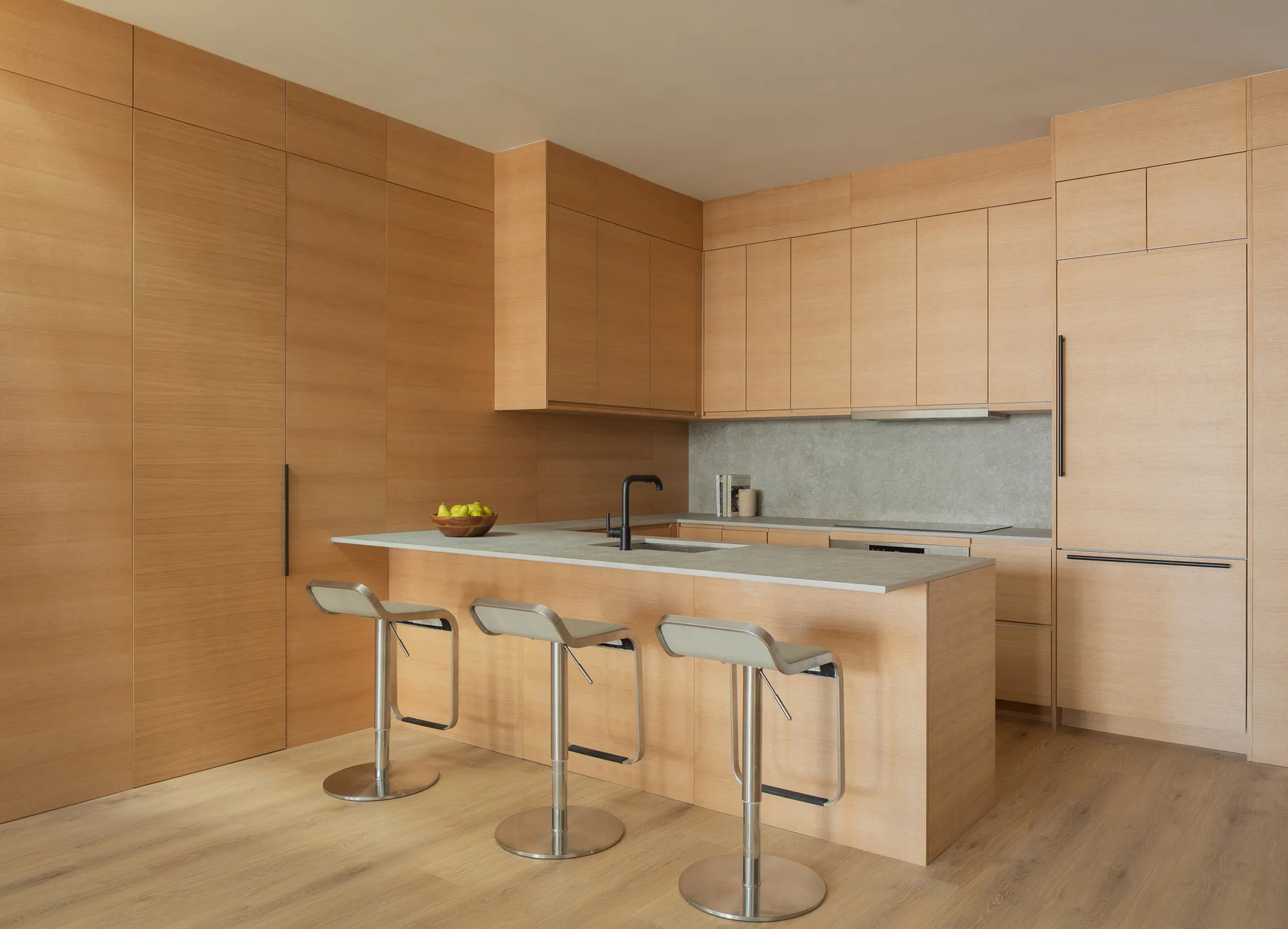 A minimalist kitchen features full-height light wood cabinetry, a matching wood kitchen island with a grey countertop, and three metal bar stools.