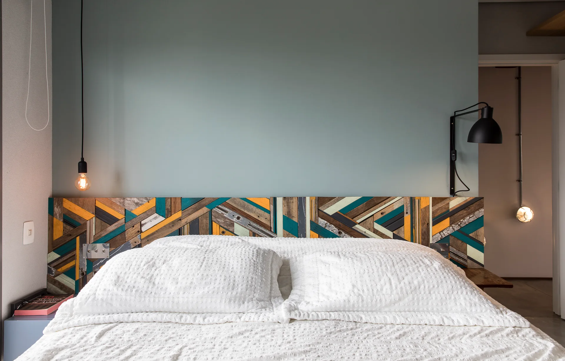 A bed with white textured bedding features a colorful geometric wood mosaic headboard set against a matte blue wall with asymmetrical bedside lamps.