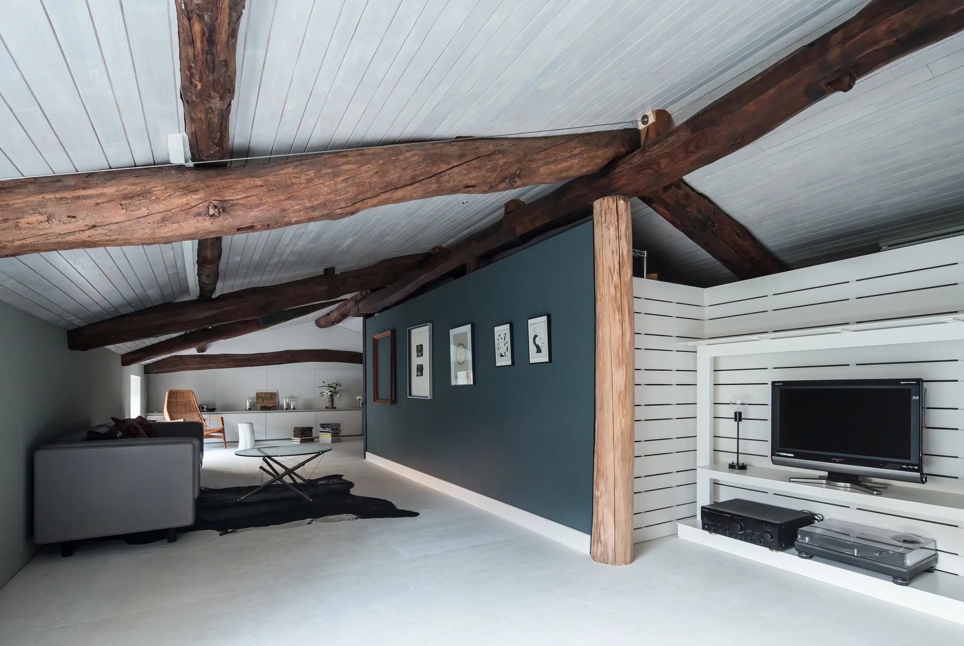 A loft living room features exposed dark wood ceiling beams, a white plank ceiling, a dark blue partition wall, and a white media unit with a television.