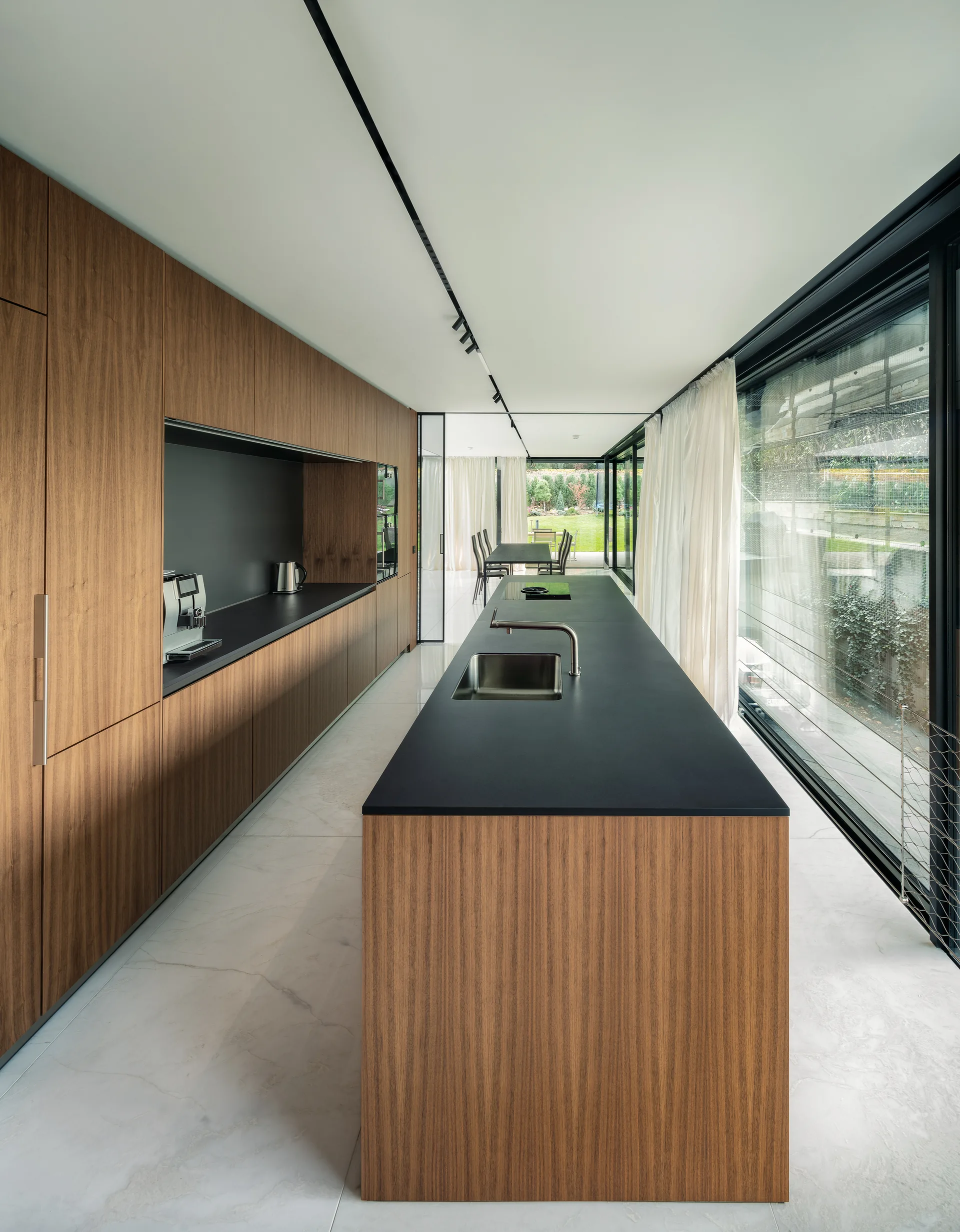 A modern kitchen featuring full-height wood veneer cabinets, a long wood island with a dark countertop, and large glass walls with sheer curtains.