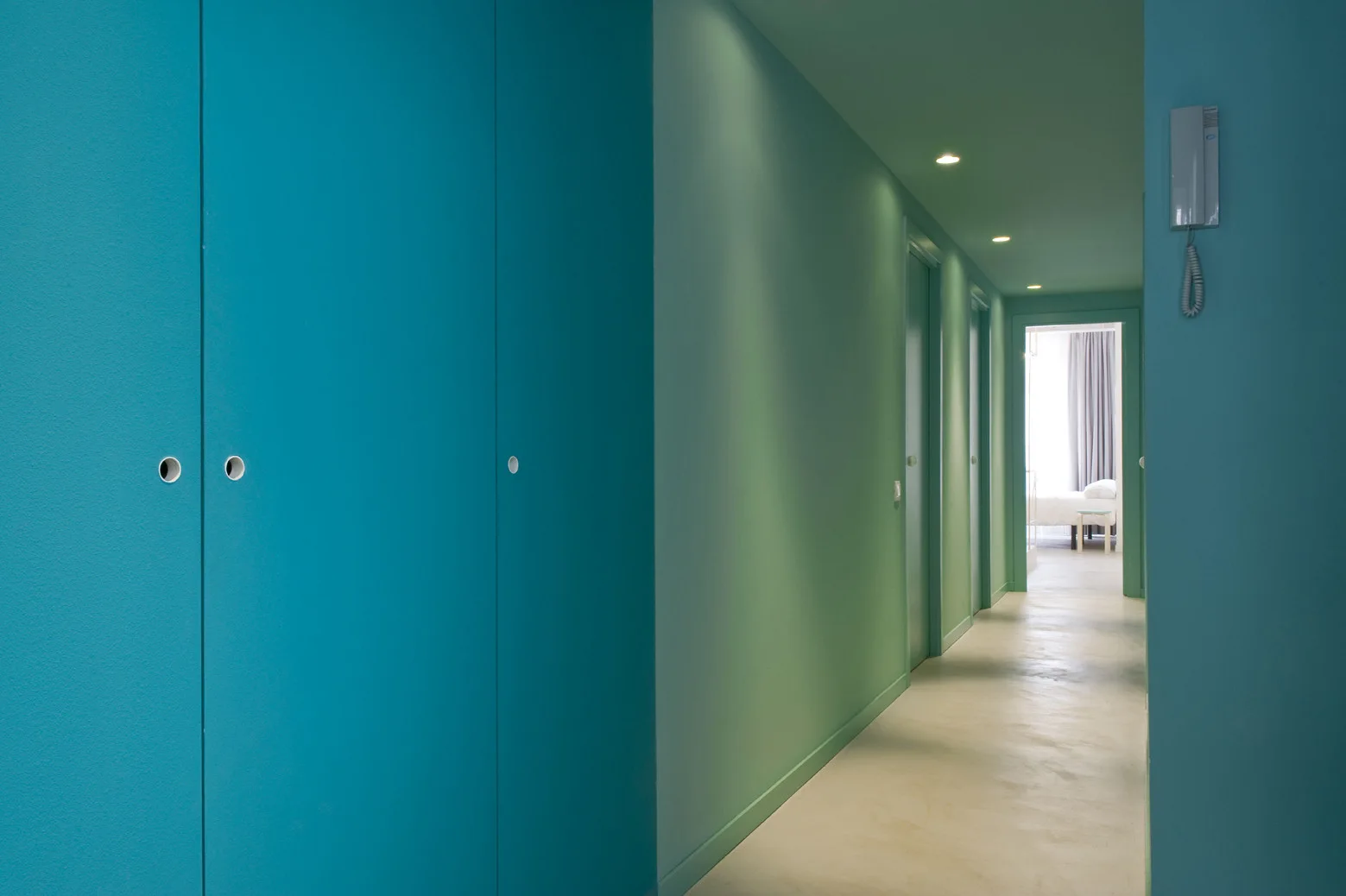 A long minimalist hallway features vibrant teal walls, matching painted ceiling and cabinet doors, and a smooth off-white floor lit by recessed lights.