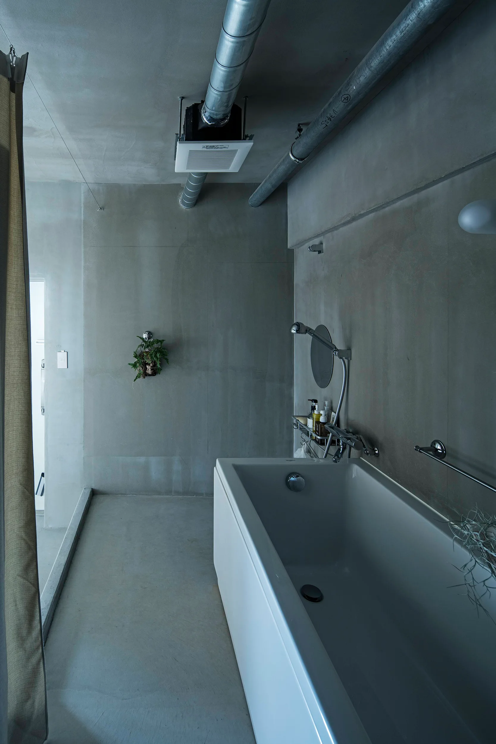 A minimalist bathroom features raw concrete walls and floors, exposed ceiling pipes, and a modern white rectangular bathtub.