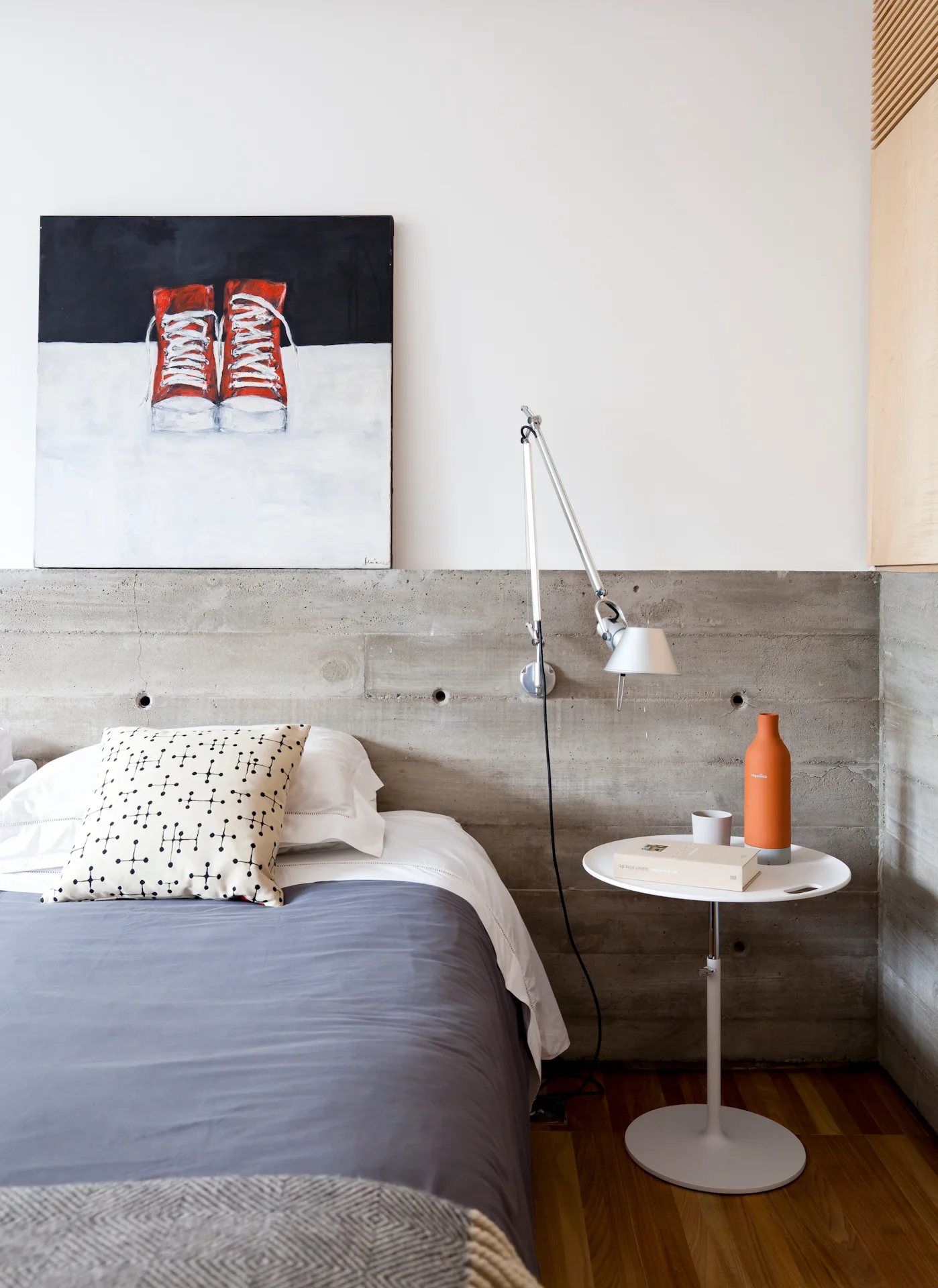 A minimalist bedroom featuring a bed against an exposed concrete wall, a white pedestal side table, and a contemporary painting of red sneakers.
