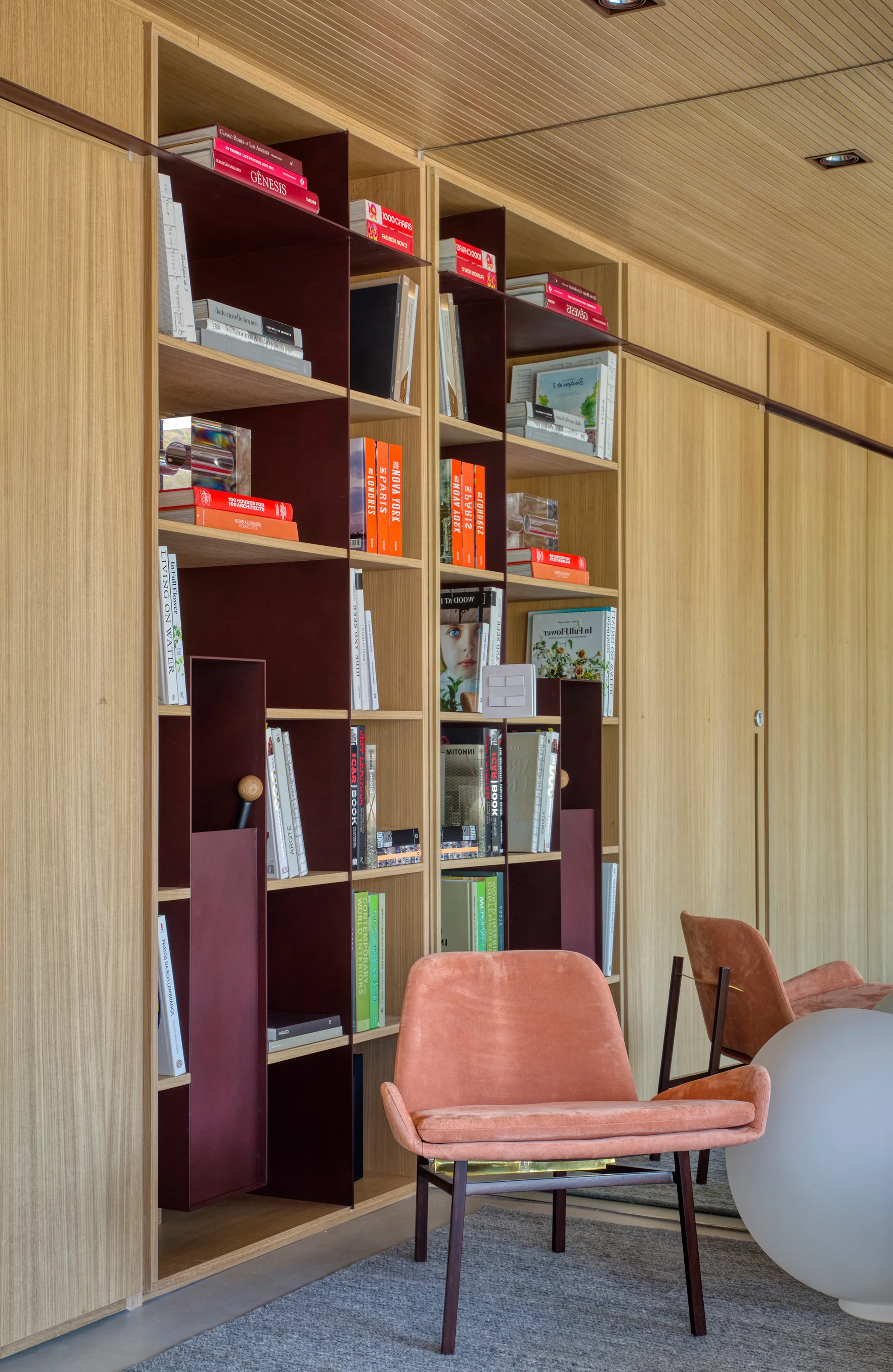 A light wood built-in bookcase features deep burgundy partitions next to a coral velvet armchair on a grey rug under a wood planked ceiling.