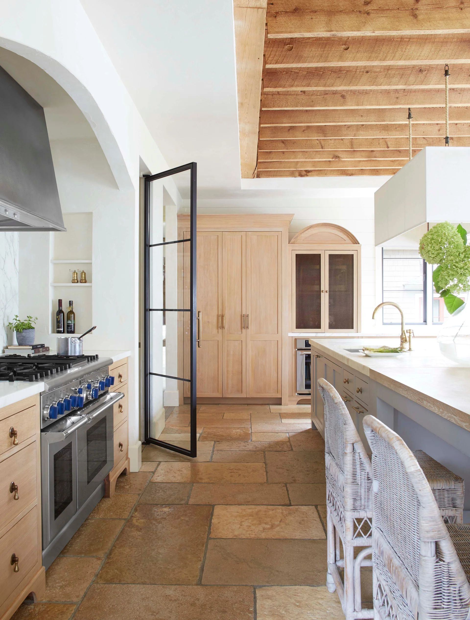 A transitional kitchen features natural stone tile flooring, a planked wood ceiling, custom light oak cabinets, and a black-framed glass door.