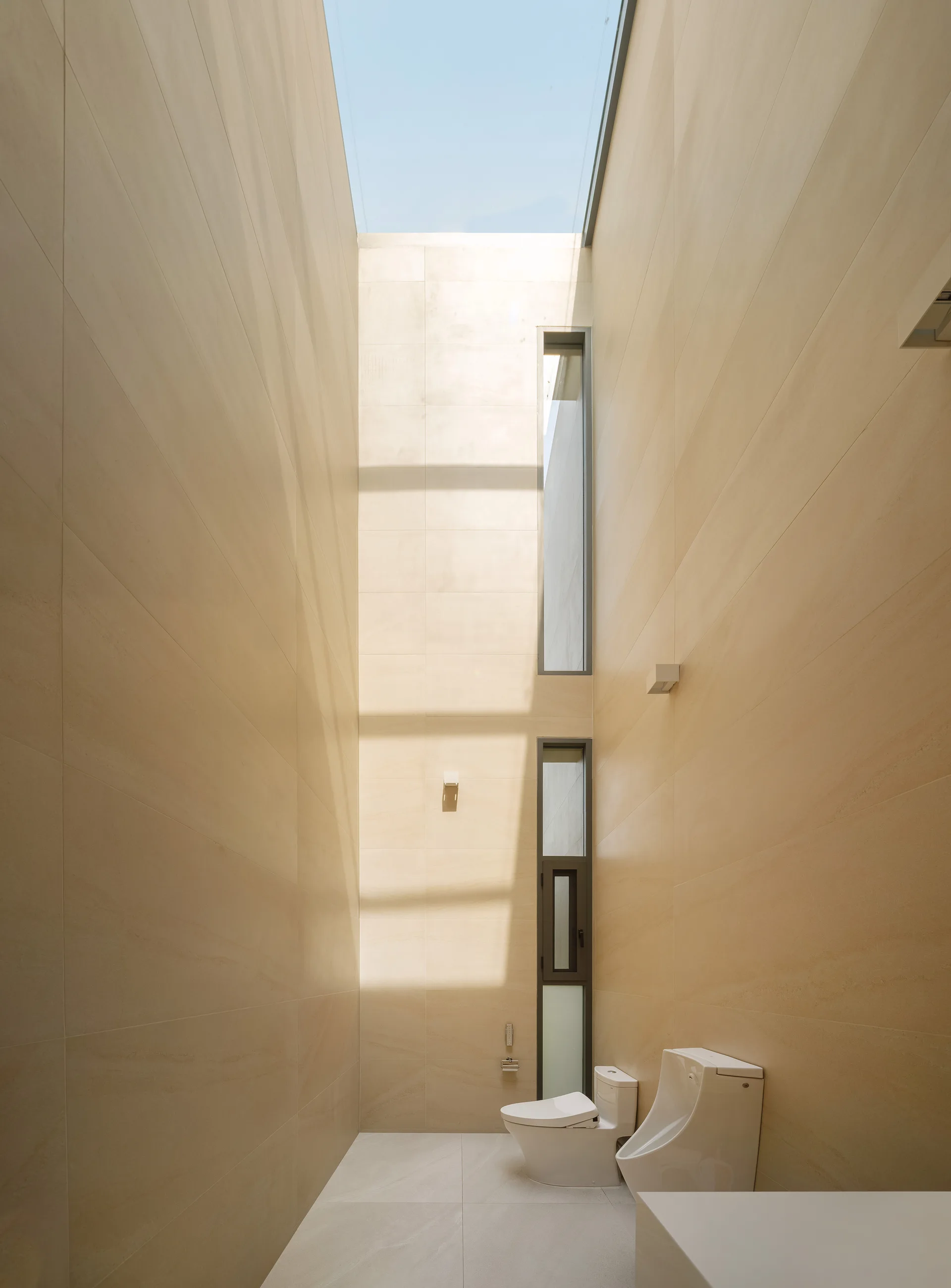 A minimalist bathroom with extremely tall beige tiled walls, an overhead skylight, and white ceramic fixtures including a toilet and urinal.