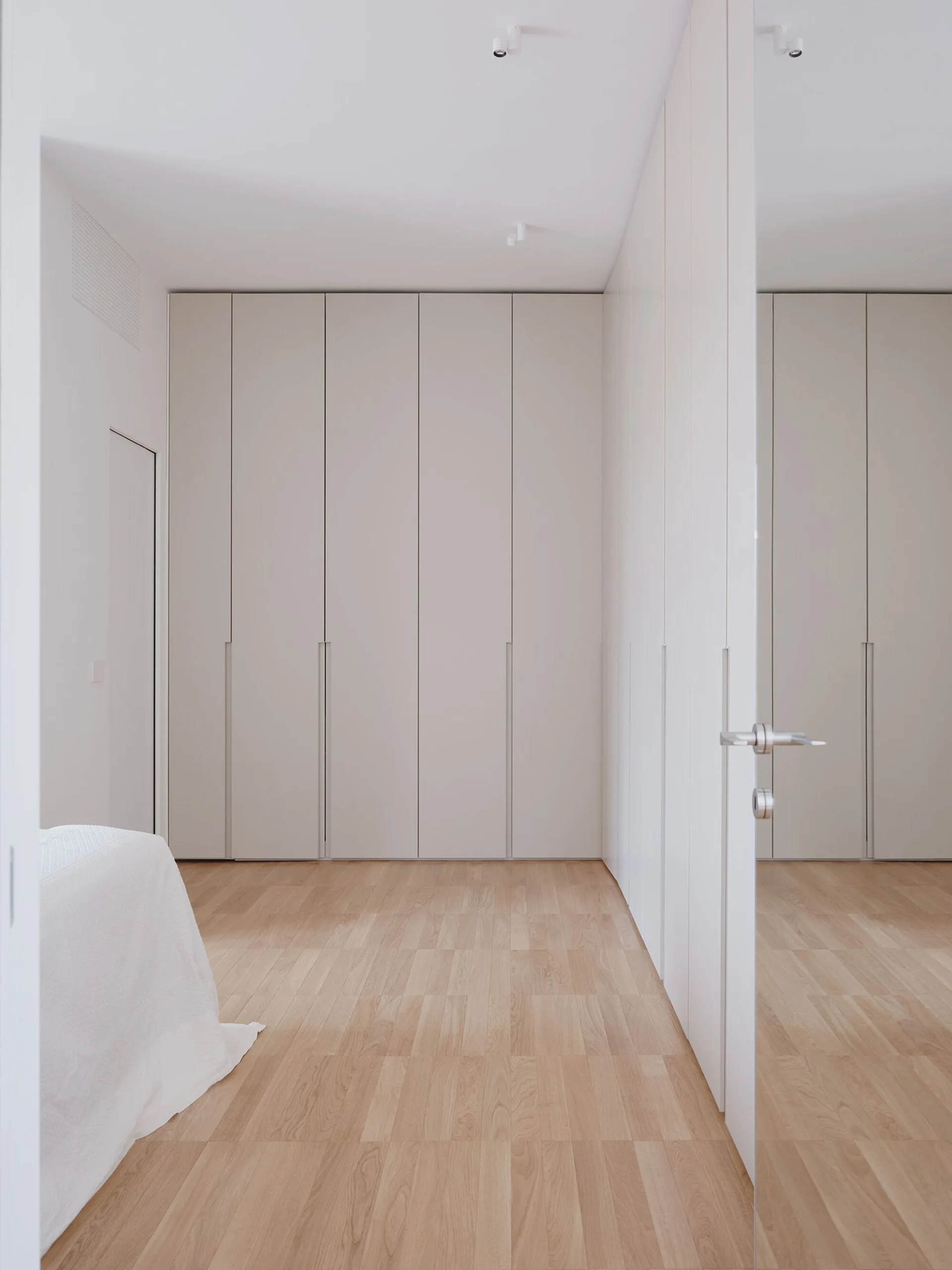 A minimalist bedroom features floor-to-ceiling white built-in wardrobes, light wood flooring, and a partially open white door.