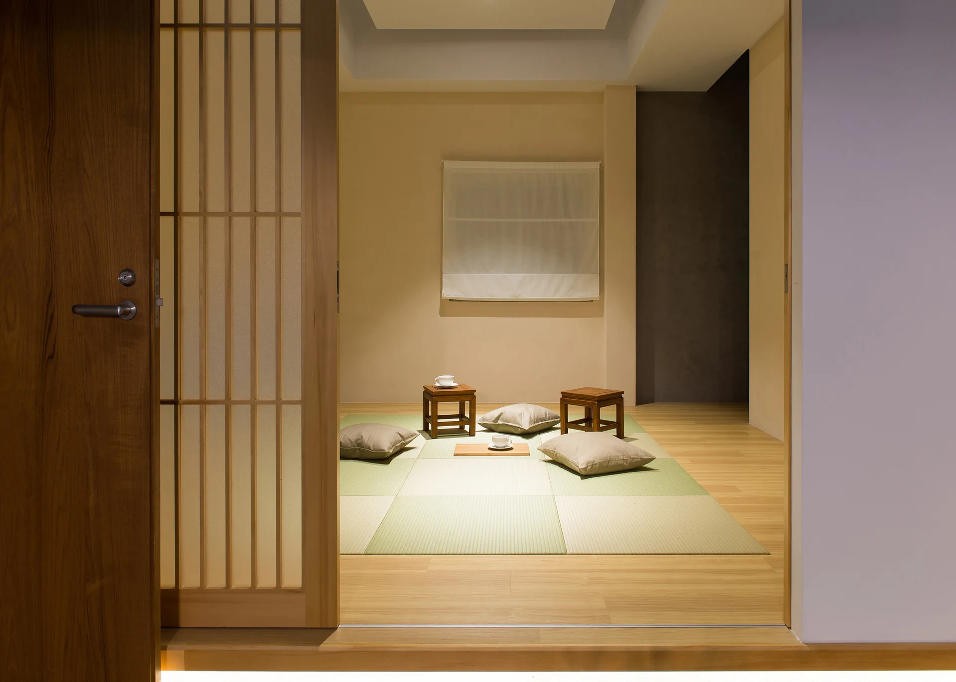A view through a wooden sliding door into a minimalist Japanese room featuring tatami mat flooring, low wooden tables, and floor cushions.