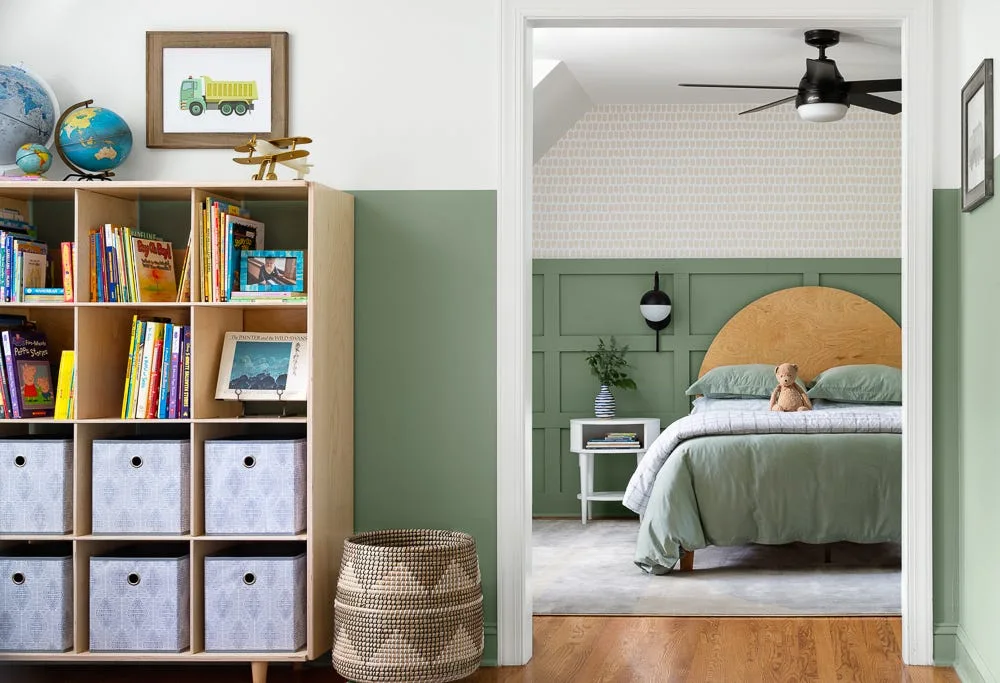 A doorway views a child's bedroom featuring sage green wainscoting, an arched wooden headboard, and a light wood toy storage bookcase.