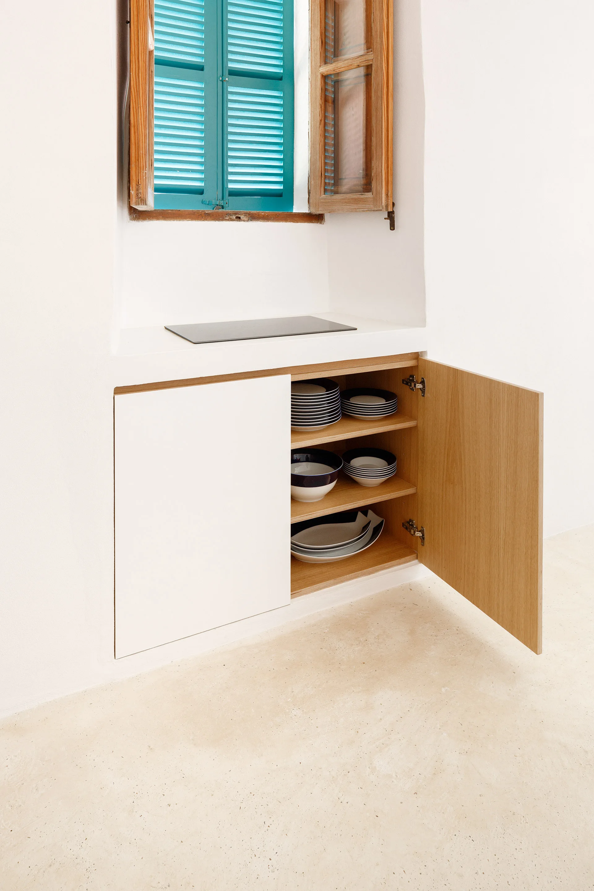 A built-in white plaster kitchen cabinet with an open wooden door sits below a recessed window with bright turquoise blue wooden shutters.