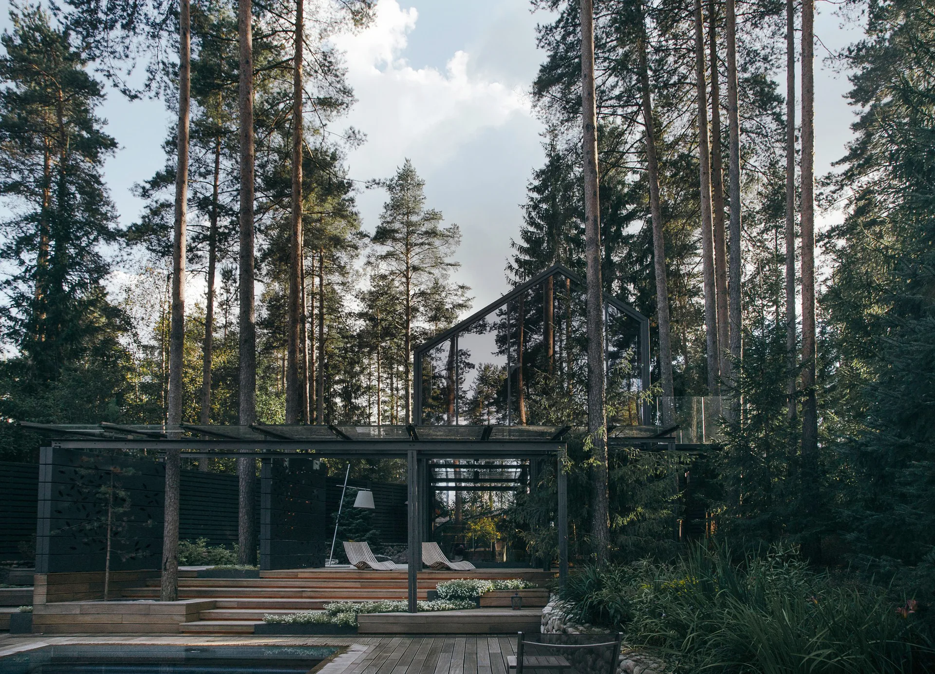 A terraced wooden deck with lounge chairs and a pool sits surrounded by tall pine trees beneath a modern glass and metal canopy structure.