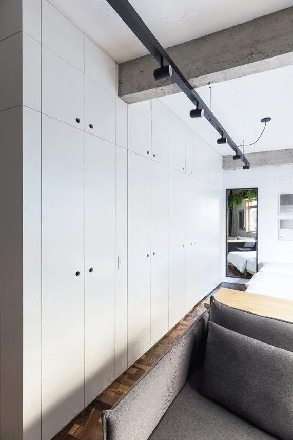 A minimalist bedroom featuring integrated white built-in wardrobes, a concrete ceiling with black track lighting, and herringbone parquet floors.