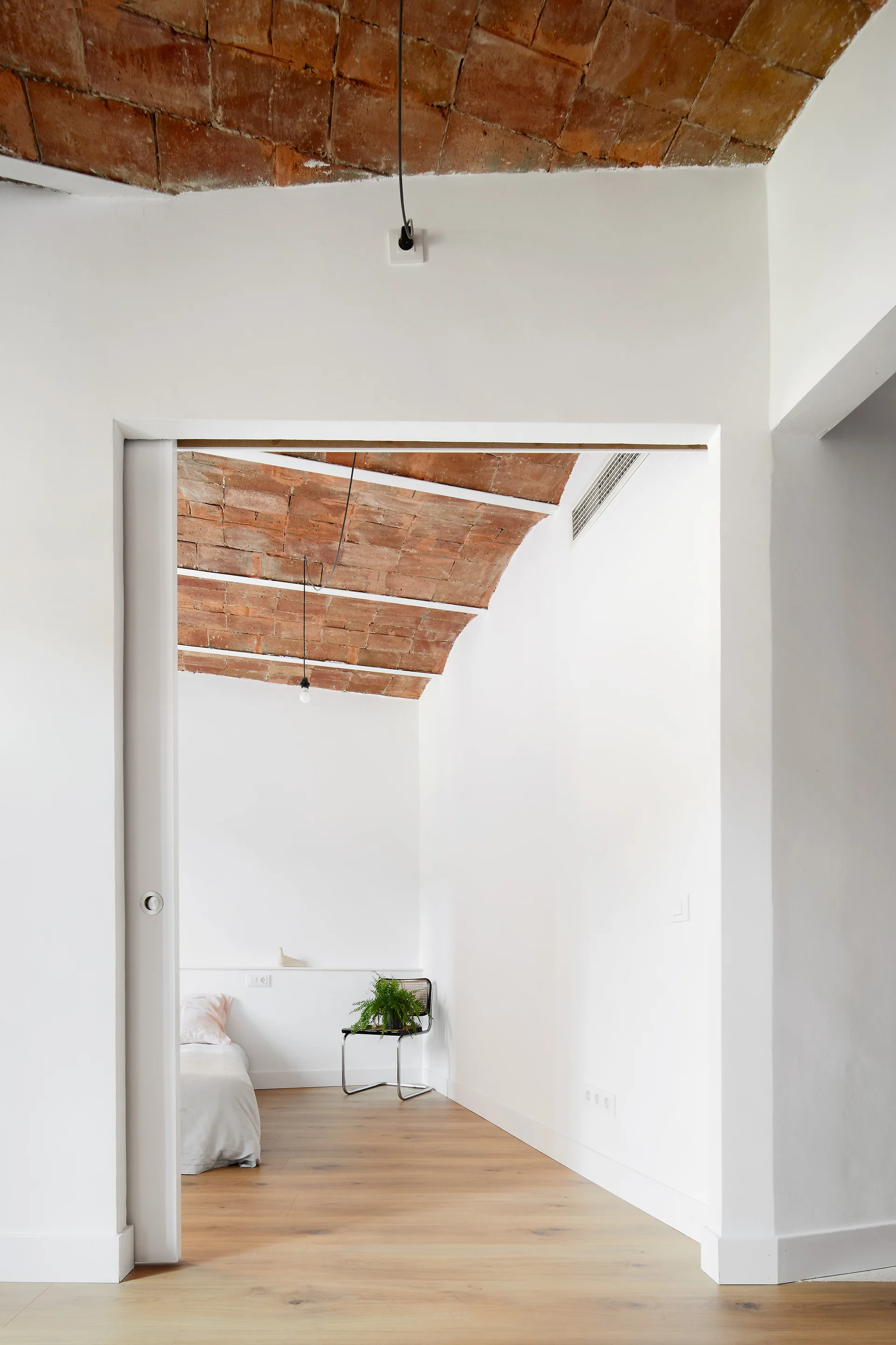 View through a wide doorway into a minimalist bedroom with a curved terracotta brick ceiling and light wood floors.