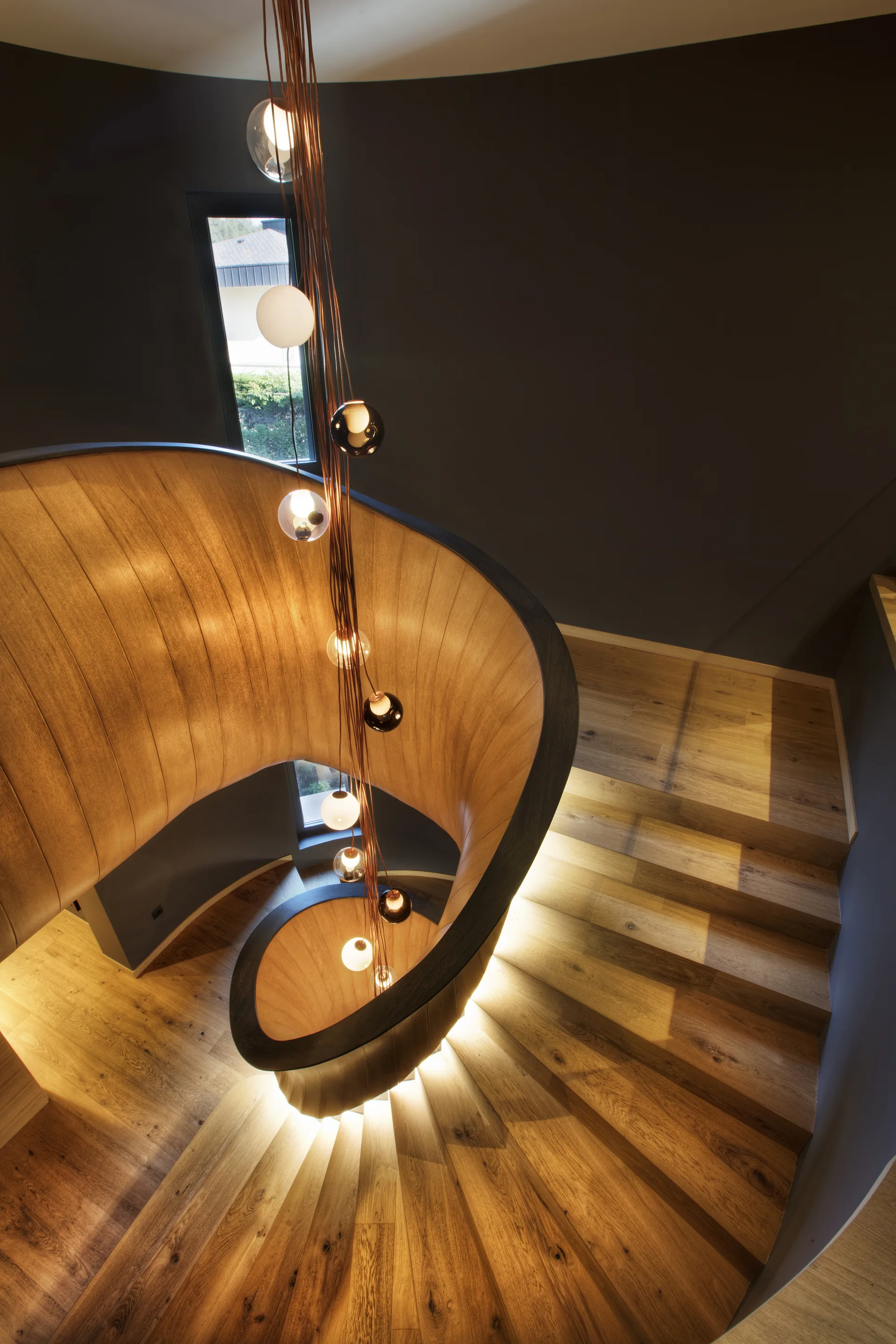 A top-down view of a curved wooden spiral staircase featuring warm integrated step lighting and a cascading multi-bulb pendant chandelier.