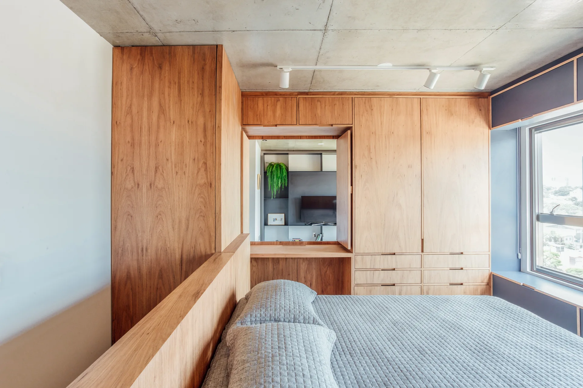 A minimalist bedroom featuring grey quilted bedding next to a large built-in wooden wardrobe and desk beneath an exposed concrete ceiling.