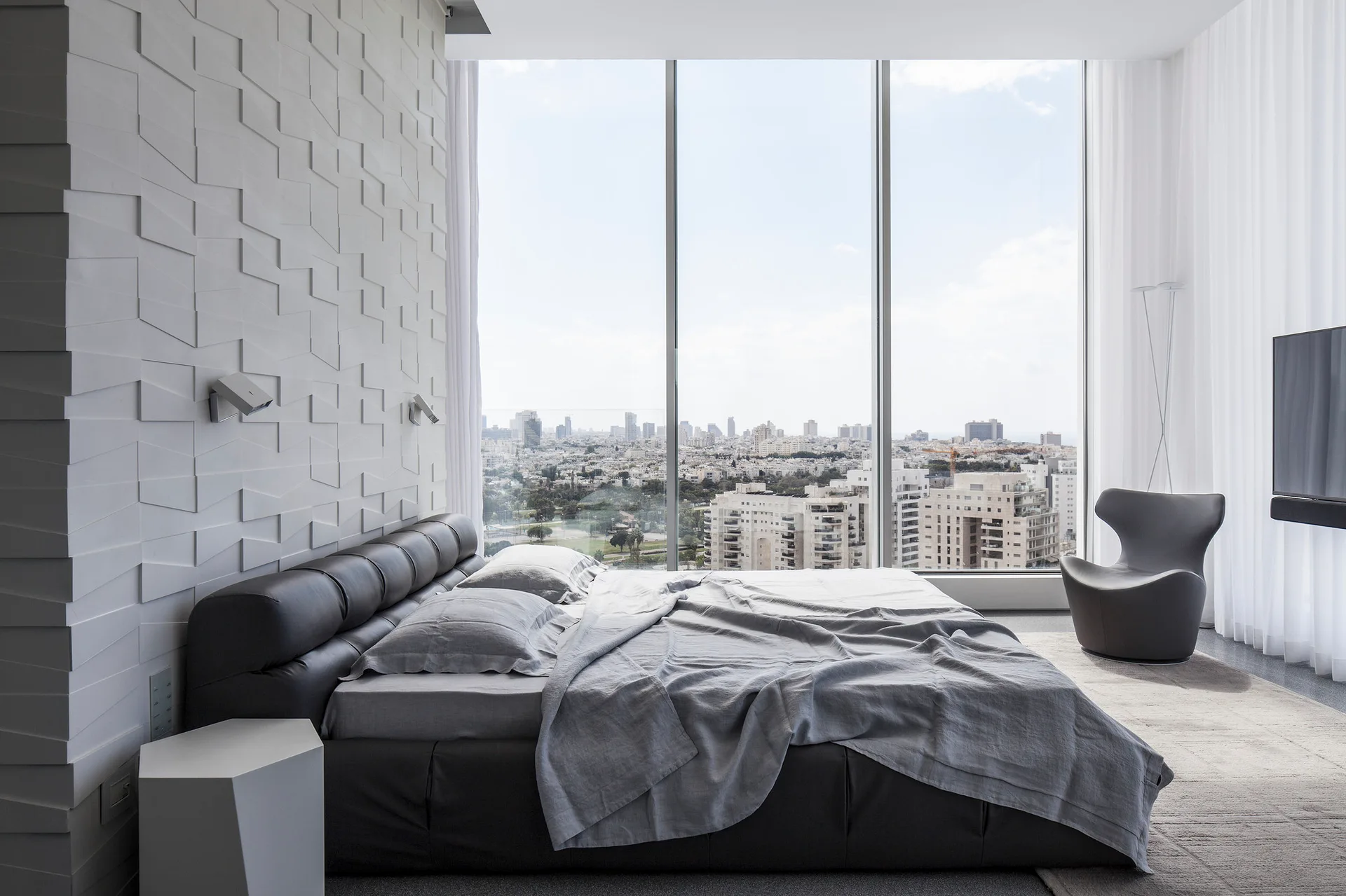 A minimalist bedroom with a charcoal upholstered bed, a white geometric textured wall, and floor-to-ceiling windows overlooking a city skyline.