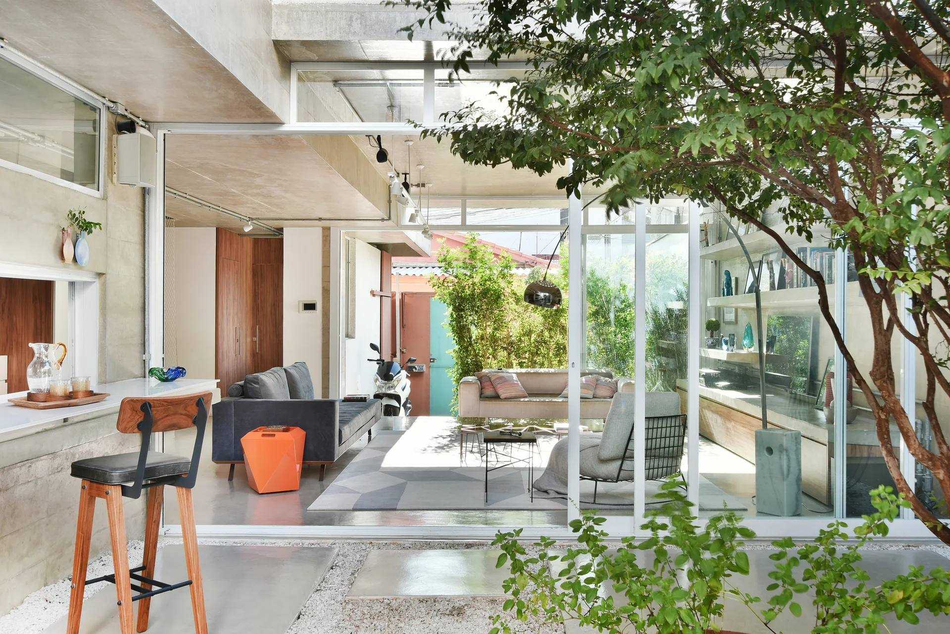 An indoor outdoor concrete living room with a gray sofa and wood console seen through open sliding glass doors next to a courtyard tree.