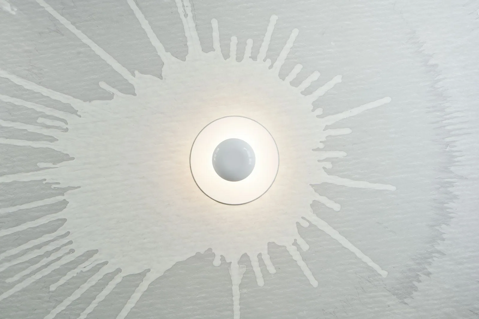 A circular white wall sconce glowing with diffused light is centered on a grey plaster wall featuring a large white paint splatter pattern.