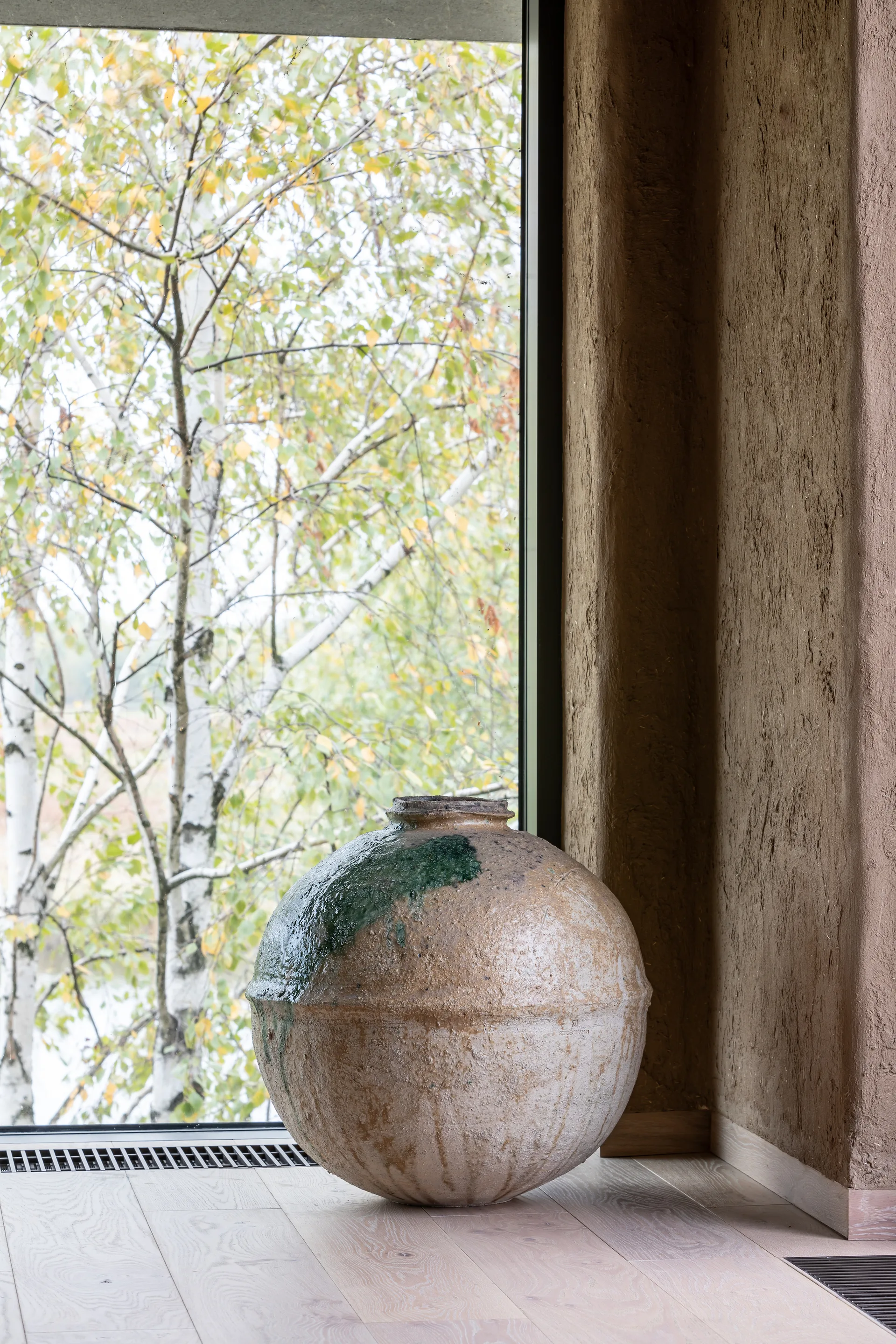 A large textured ceramic vessel rests on a light wood floor near a floor-to-ceiling window showcasing birch trees and a rough earth-toned wall.