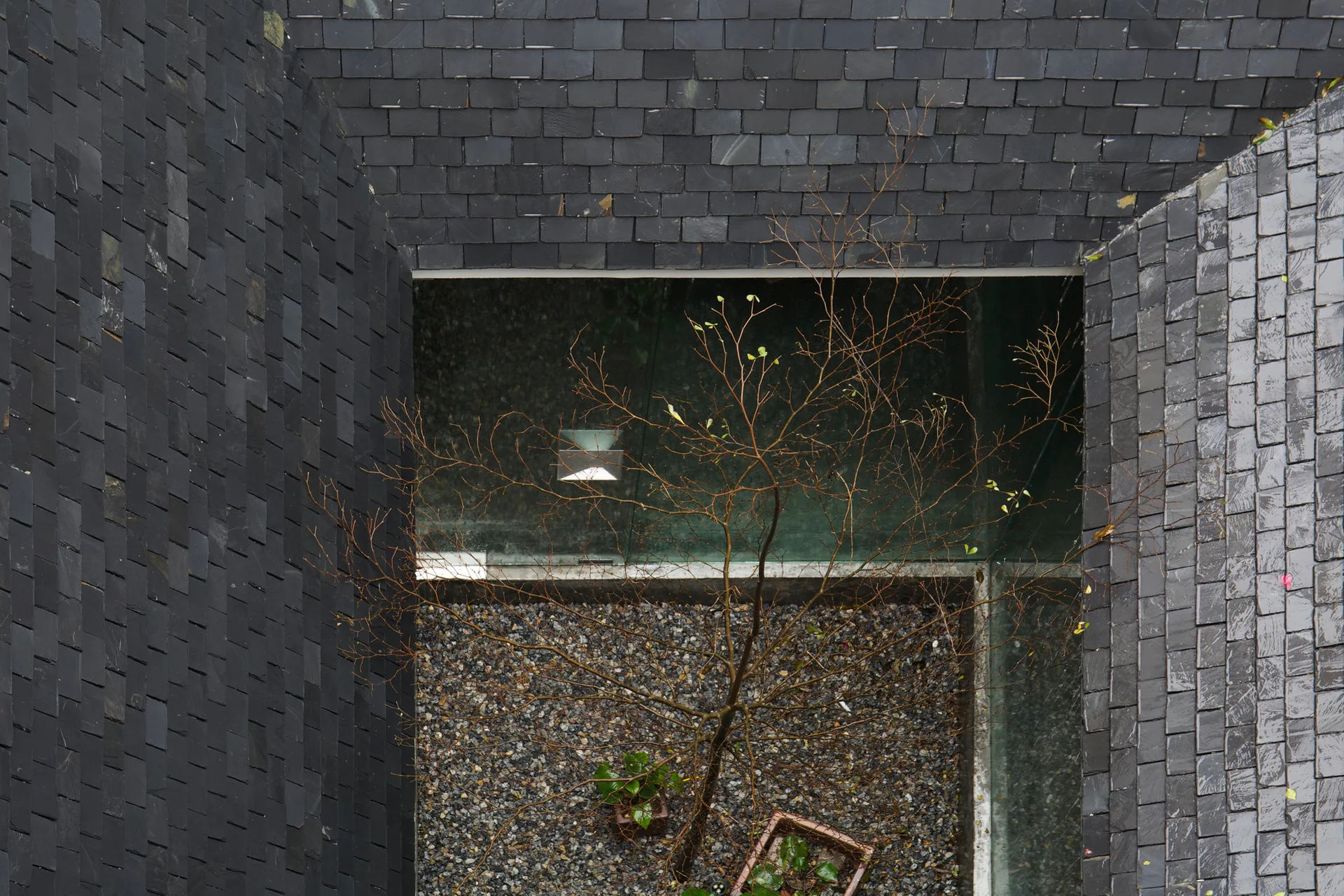 An overhead view looking down into a small square courtyard with a gravel floor and a single tree, enclosed by steep dark grey slate shingled walls.