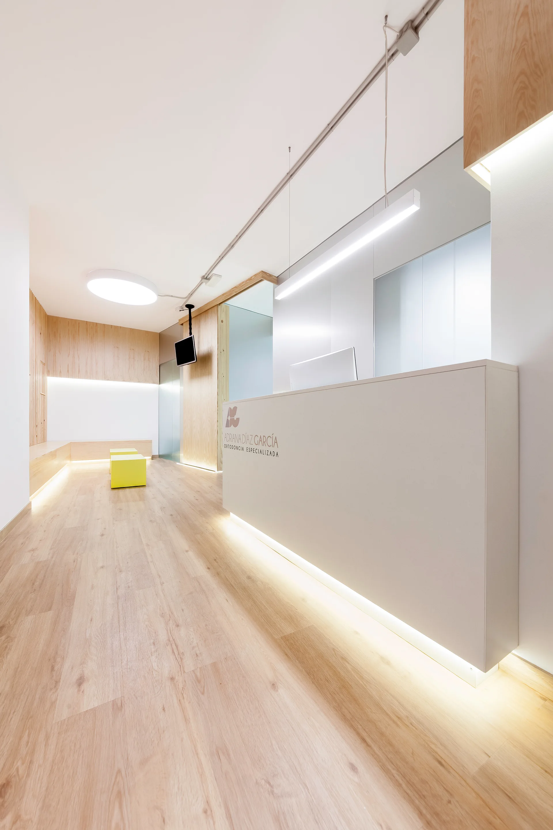 A minimalist reception area features a large white desk with under-counter lighting, light wood floors, and warm wood wall panels.