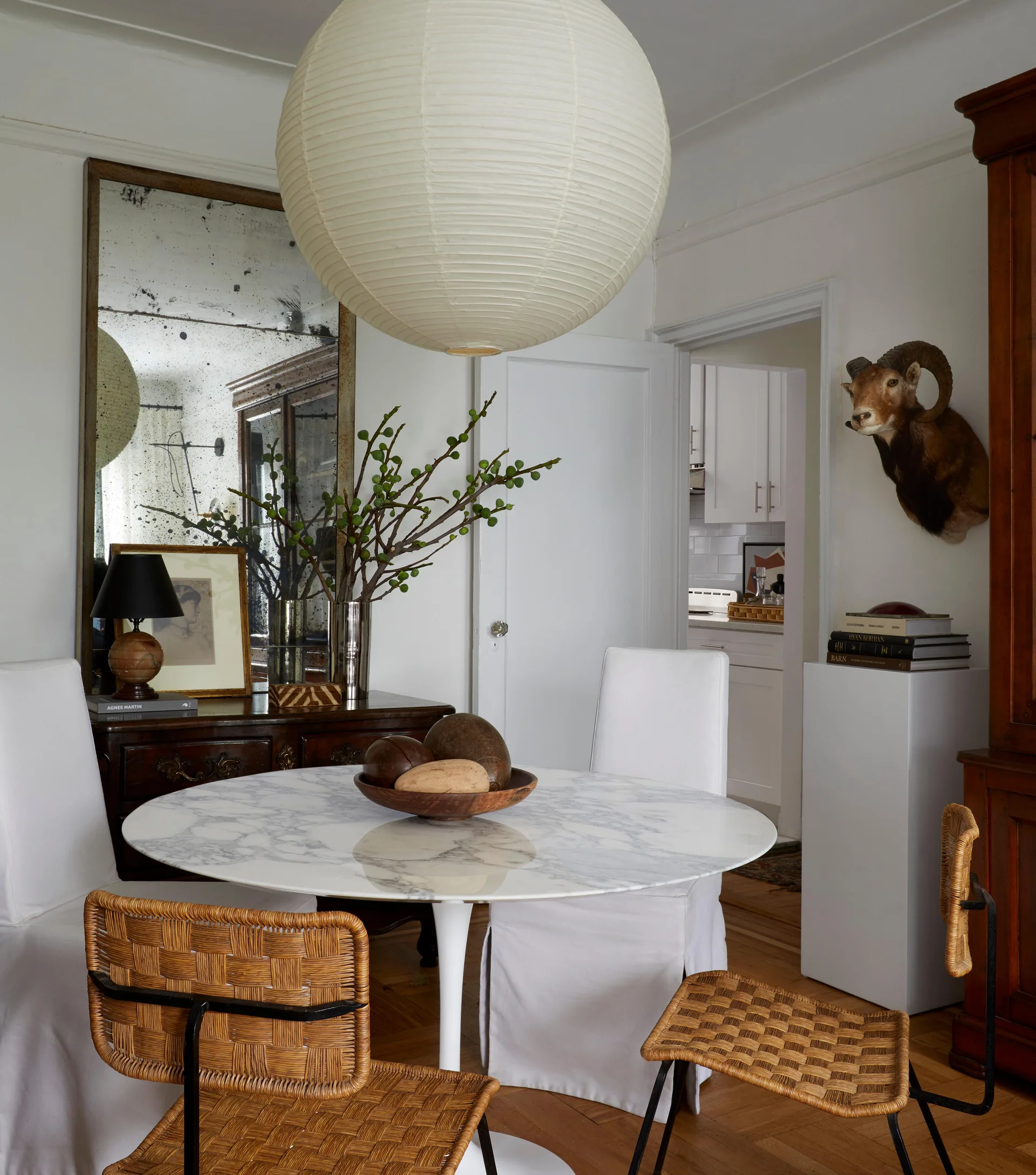 A round marble dining table is surrounded by woven rattan chairs and white slipcovered chairs under a large paper globe pendant light.