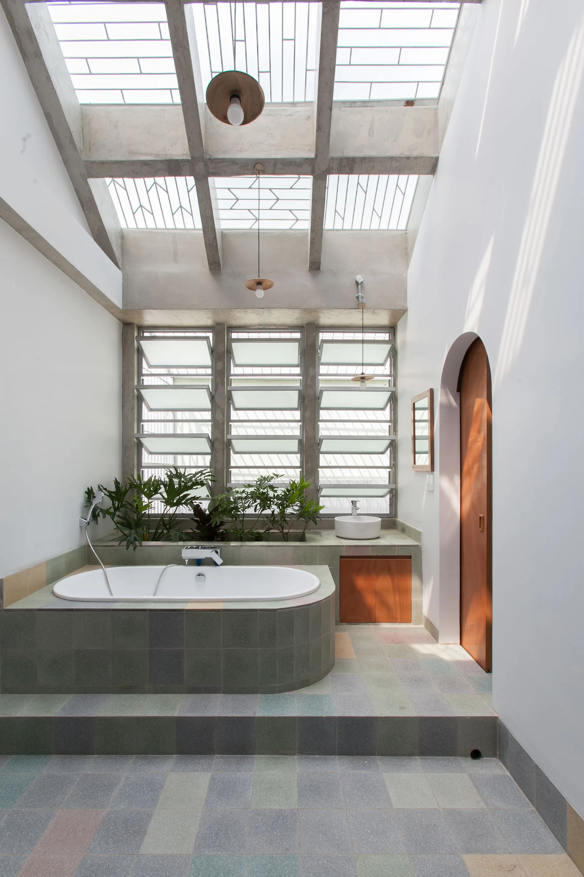 A bright bathroom with a bathtub built into a tiled deck, green plants in a planter, and a concrete grid ceiling with skylights.
