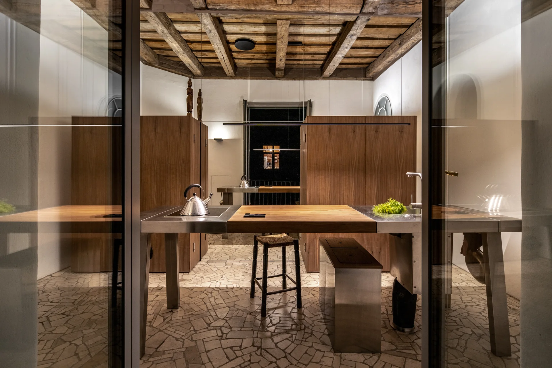 A modern kitchen featuring an island with stainless steel and wood surfaces, set beneath an exposed timber beam ceiling with a mosaic stone floor.