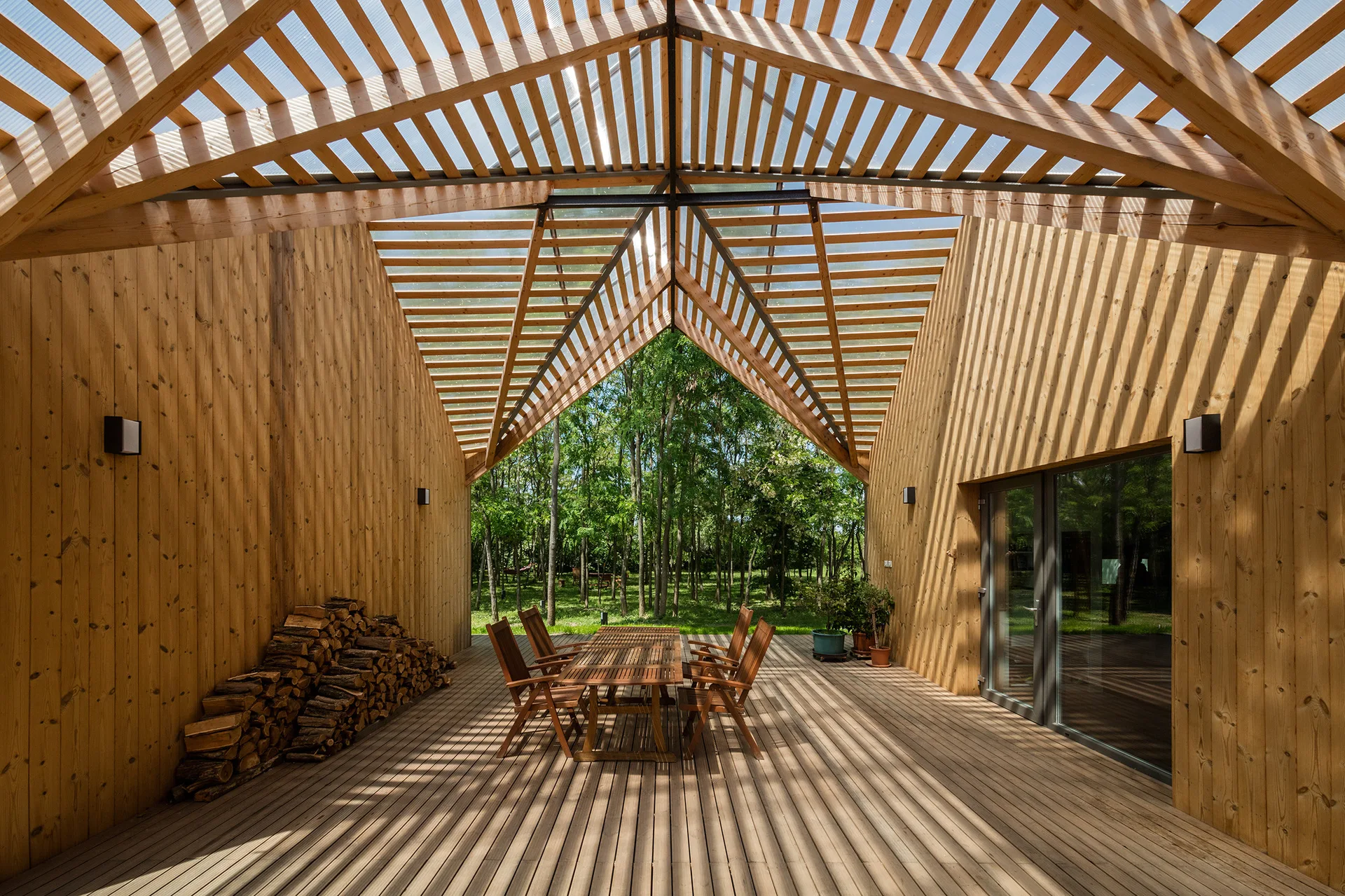 A wooden outdoor patio with a dining table and chairs set beneath a slatted gable roof that casts linear shadows on the deck.